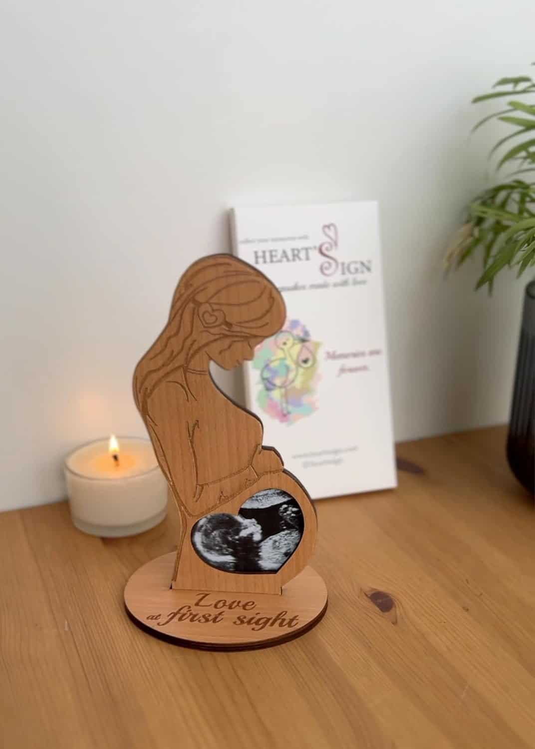 Wooden silhouette of a pregnant woman with an ultrasound image in her belly area, standing on a wooden base labeled "Love at first sight," next to a lit candle and a plant.