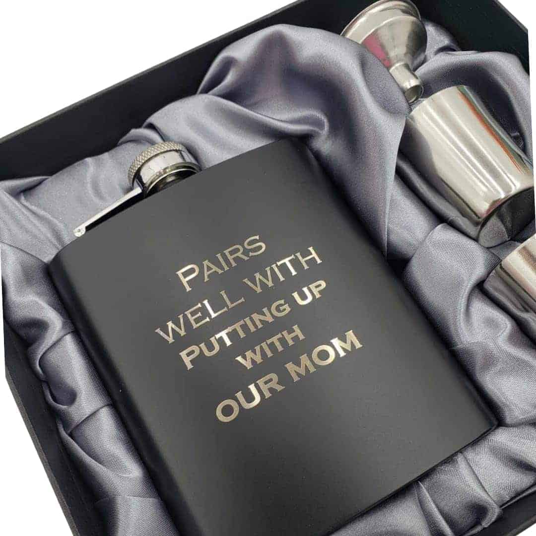 A black flask with the inscription "Pairs well with putting up with our mom" in gold text, displayed in a gift box with two metal shot glasses on gray satin fabric.