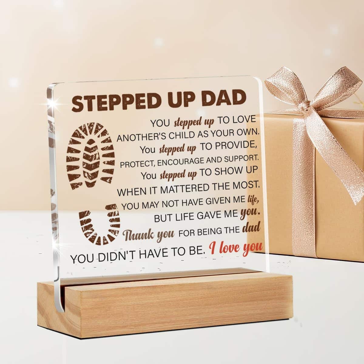 A glass plaque with a wooden base, inscribed with a message for a "stepped up dad," placed next to a gift box with a ribbon.