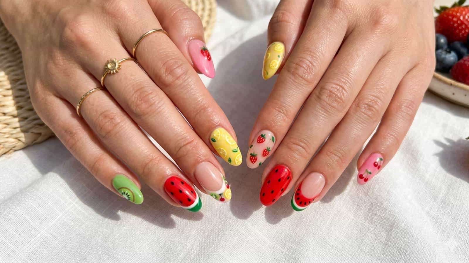 Hands with long, almond-shaped nails painted with colorful fruit designs, including watermelon, strawberry, lemon, and kiwi patterns. The hands wear gold rings and rest on a white surface.