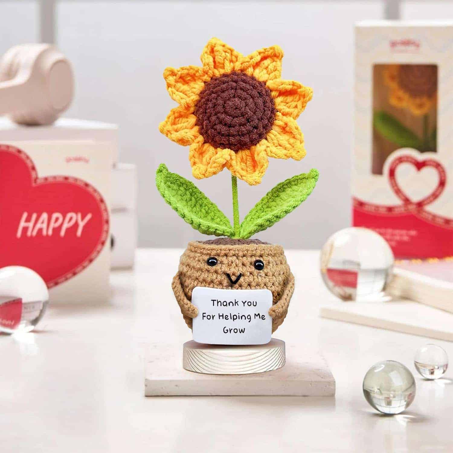A crocheted sunflower in a small pot holds a sign that says "Thank You For Helping Me Grow," surrounded by gift boxes and decorative objects on a table.