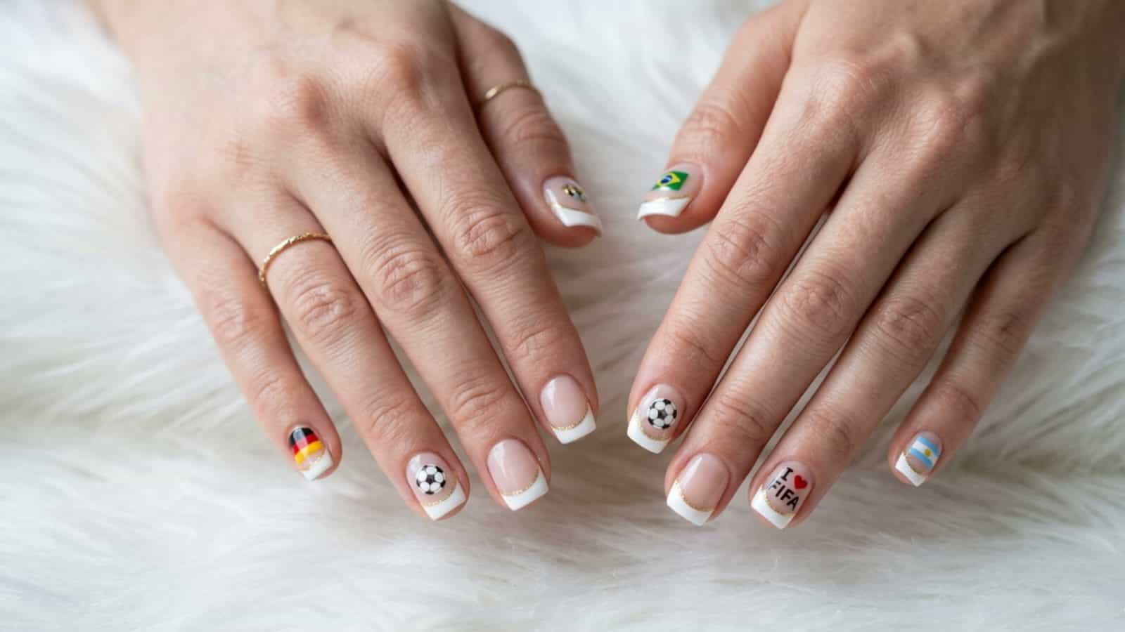 Two hands with manicured nails featuring small soccer-themed designs and flags, displayed on a white, fluffy surface.