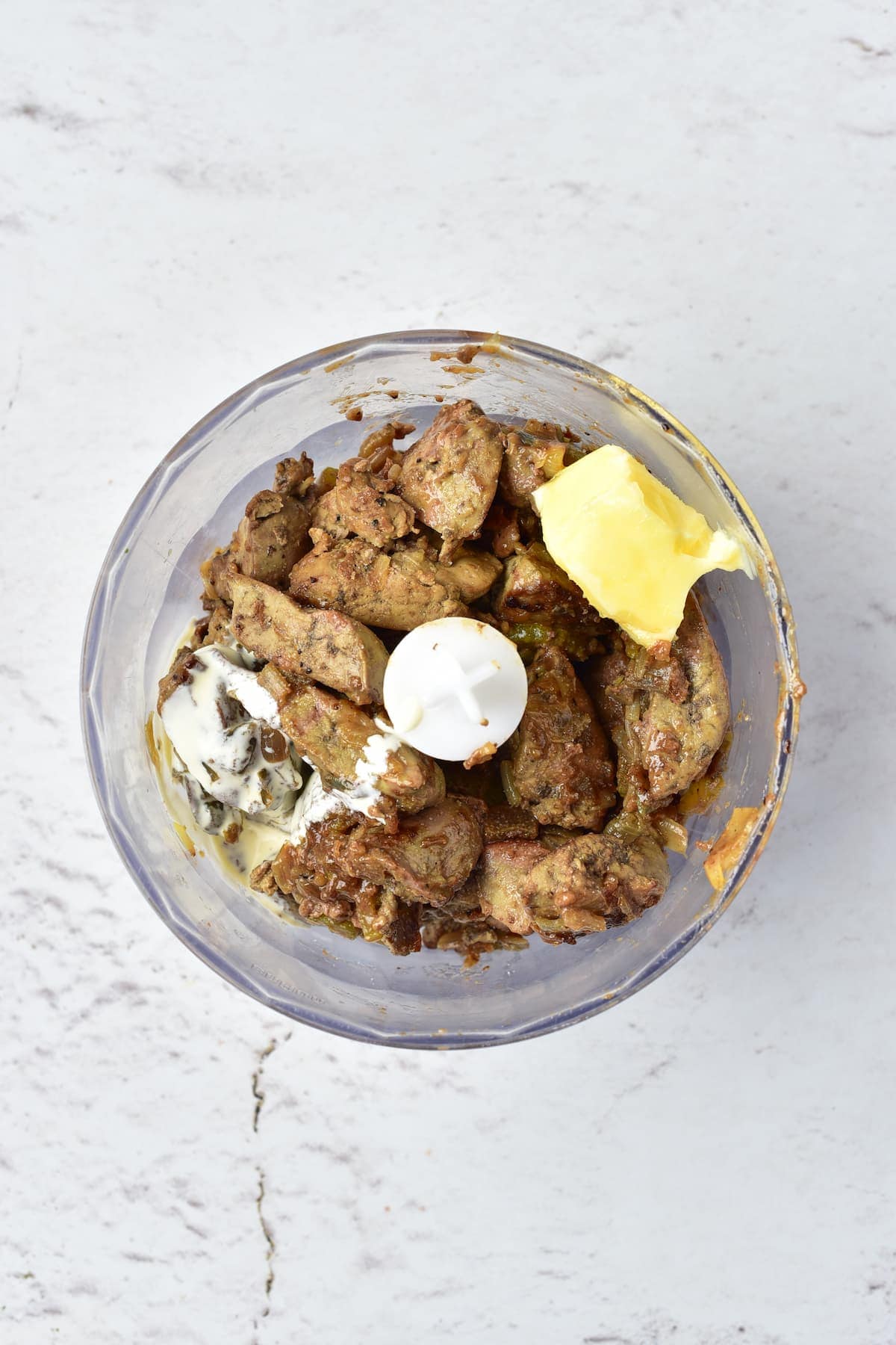 A food processor bowl containing cooked chicken liver pieces, butter, and cream cheese on a light gray surface.