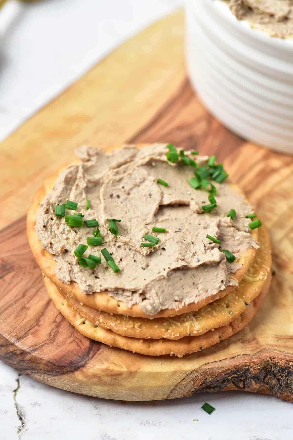 Three crackers topped with a spread and garnished with chopped chives, placed on a wooden board next to a bowl of the same spread.