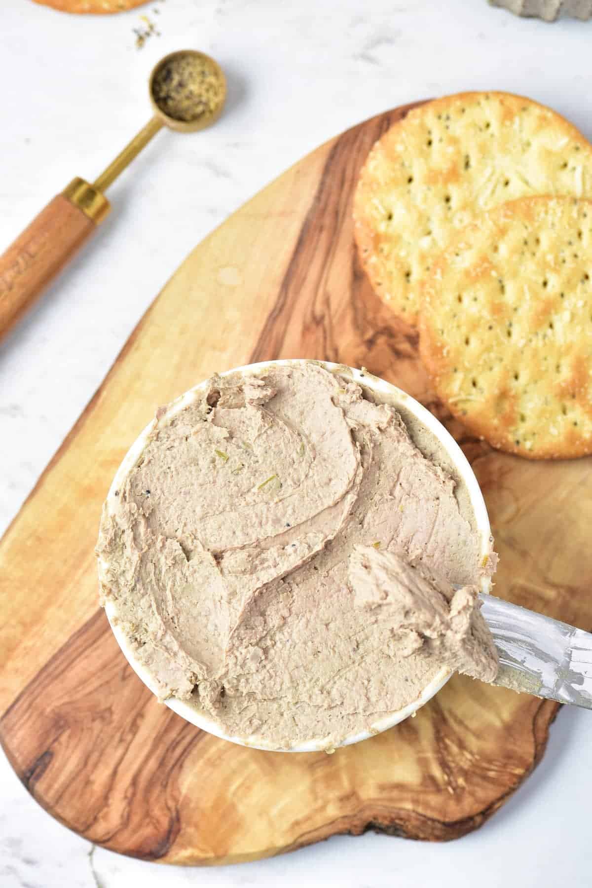 A bowl of pâté with a spreader rests on a wooden board next to round crackers.