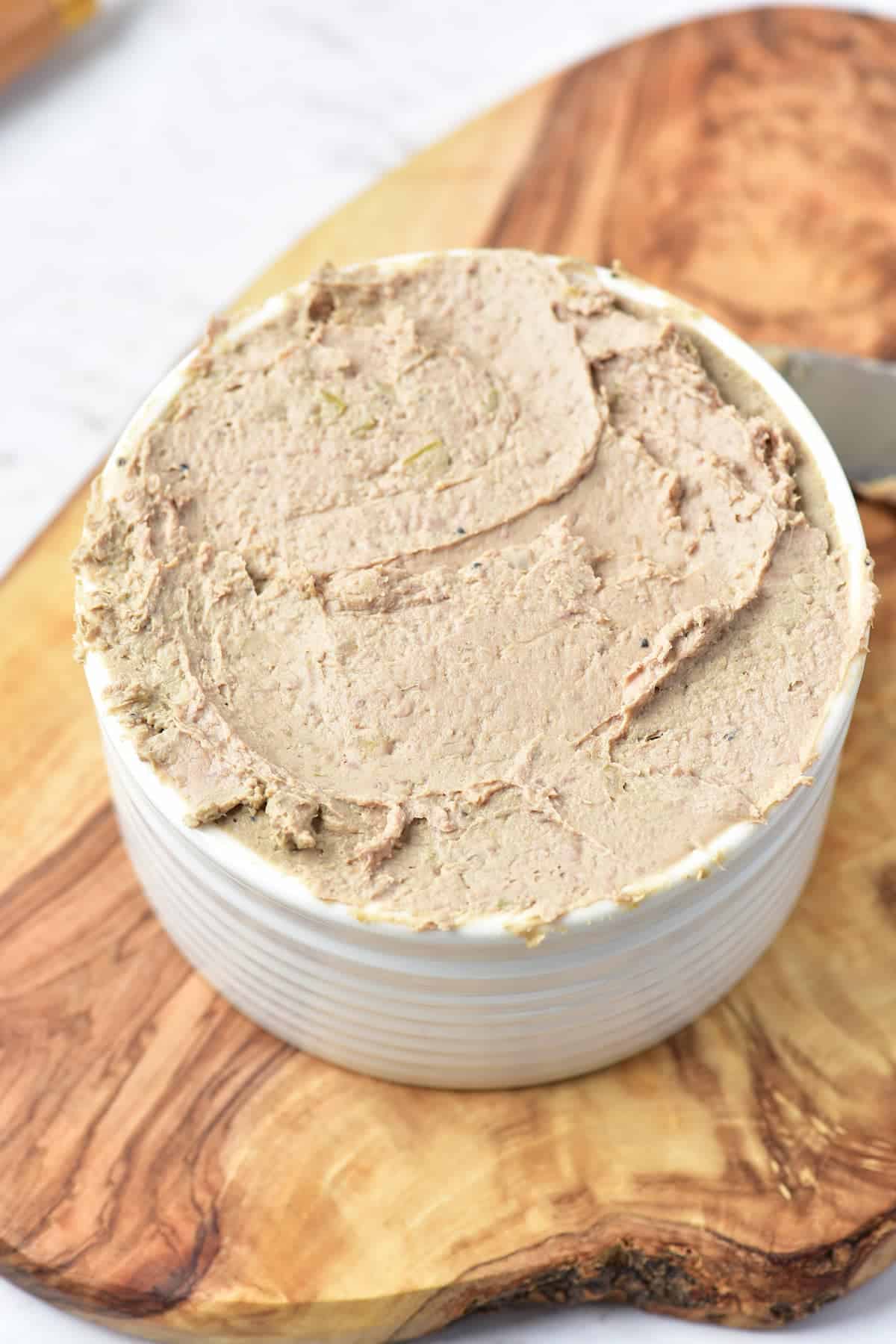 A white ramekin filled with smooth, spreadable pâté sits on a wooden cutting board.