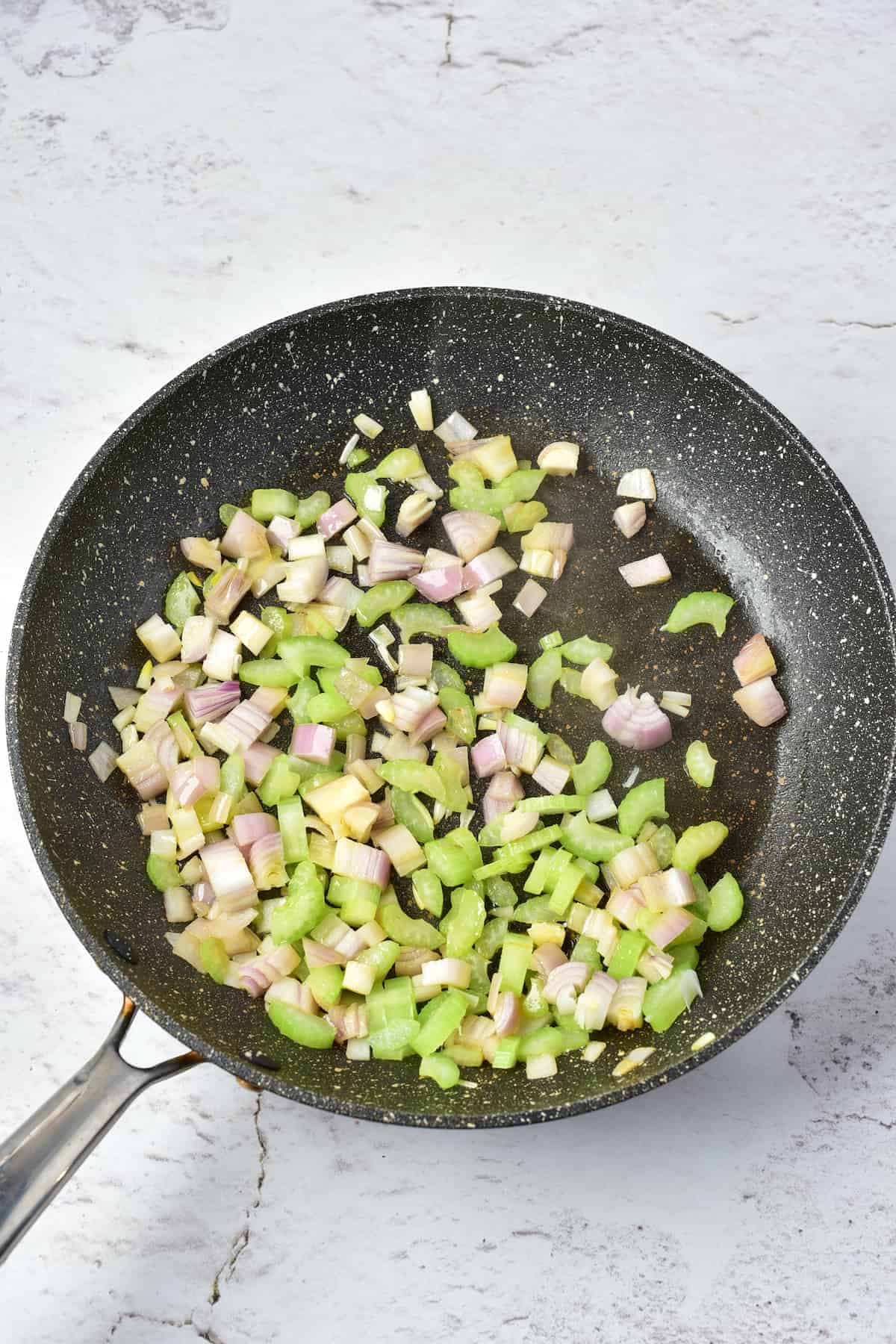 Diced onions, celery, and garlic are being sautéed in a black frying pan on a light textured surface.