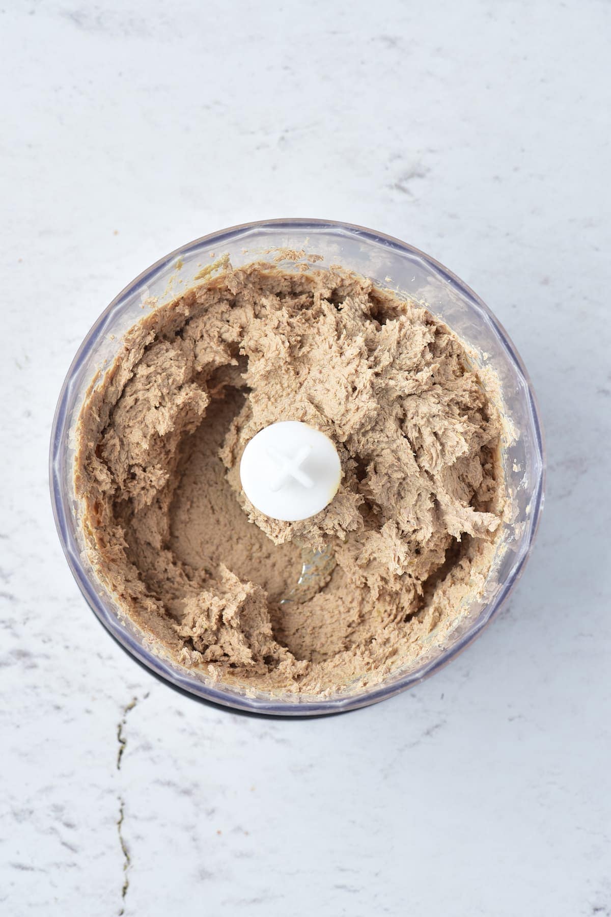 A food processor bowl filled with a blended, light brown mixture sits on a light-colored countertop.