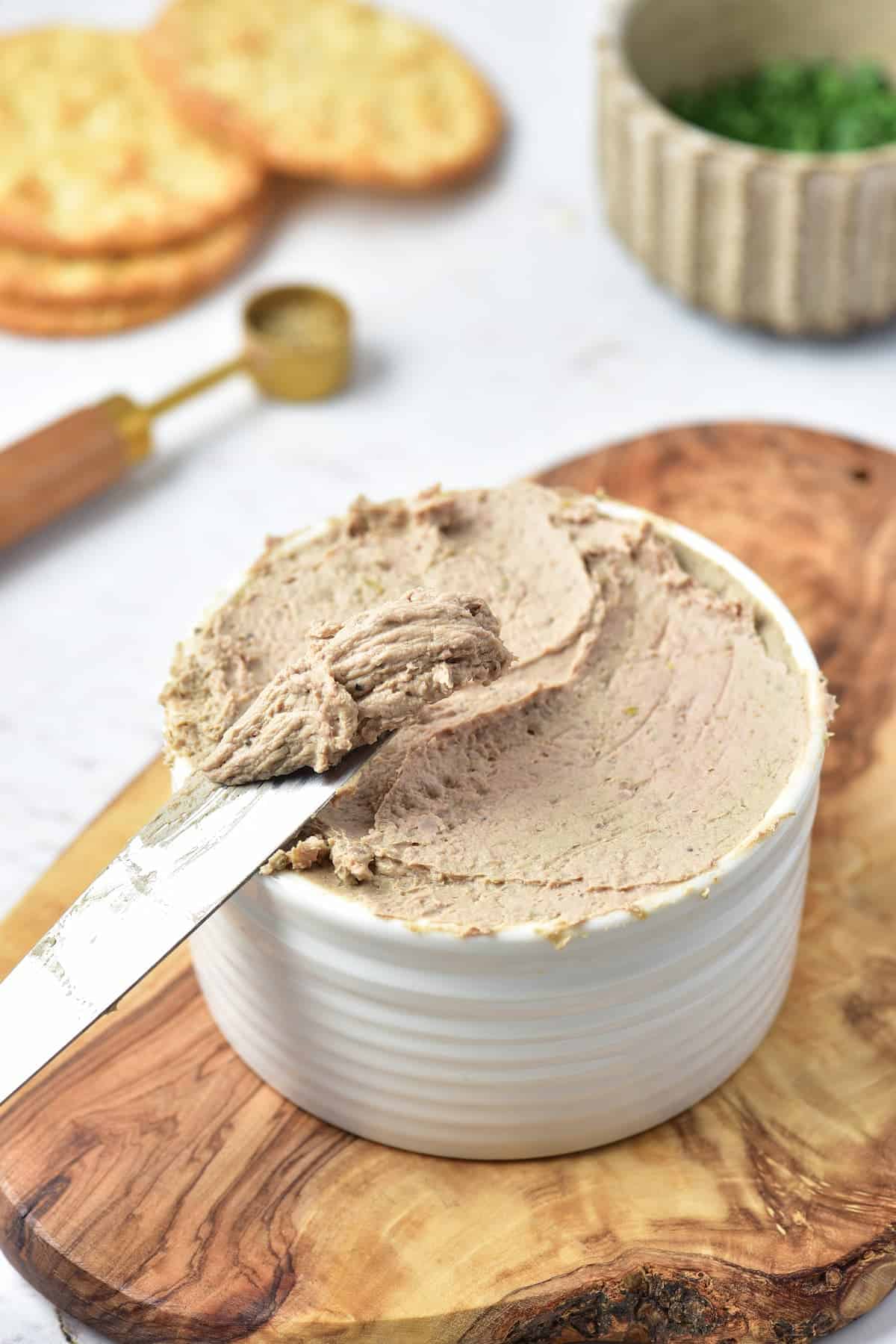 A white ramekin filled with a smooth brown spread, with a knife resting on top, sits on a wooden board. Crackers and a bowl of herbs are in the background.