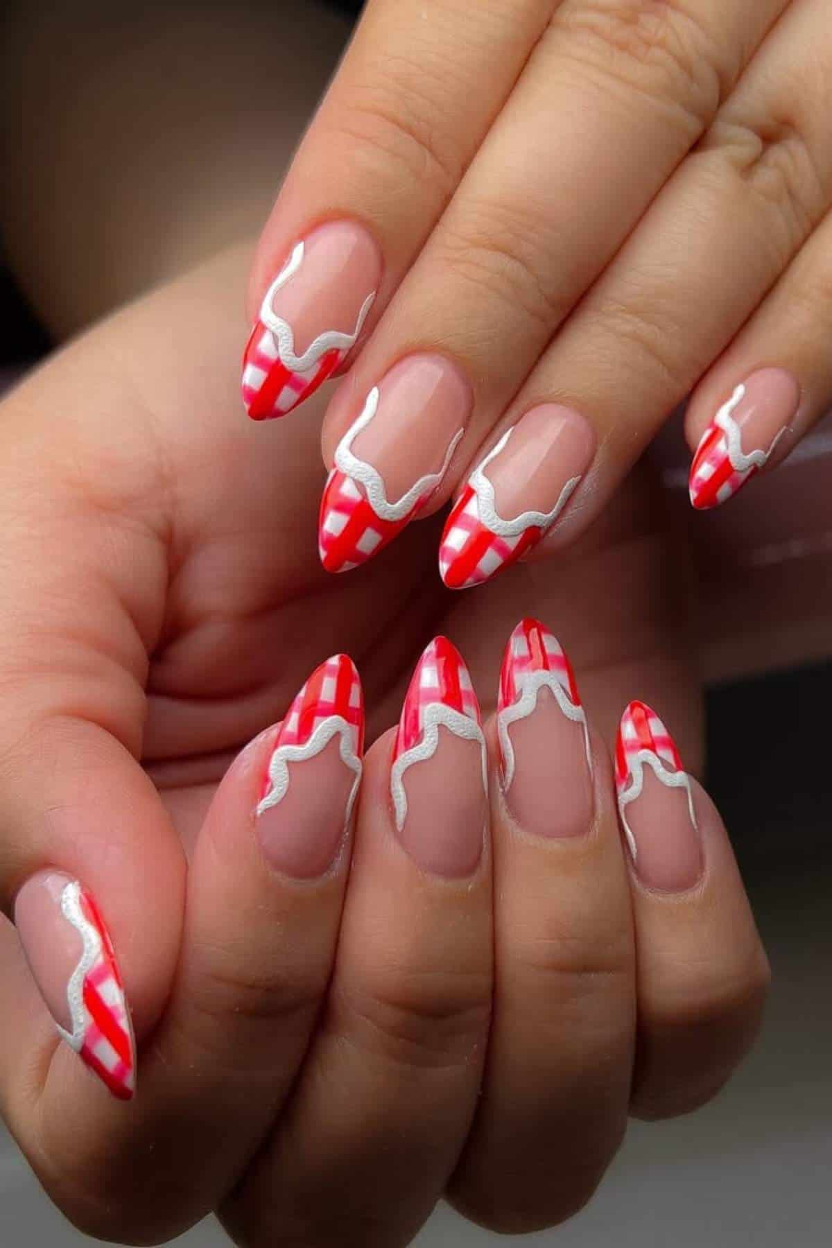 Hands with almond-shaped nails featuring a red and white checkered tip design with a wavy white outline on a natural nude base.