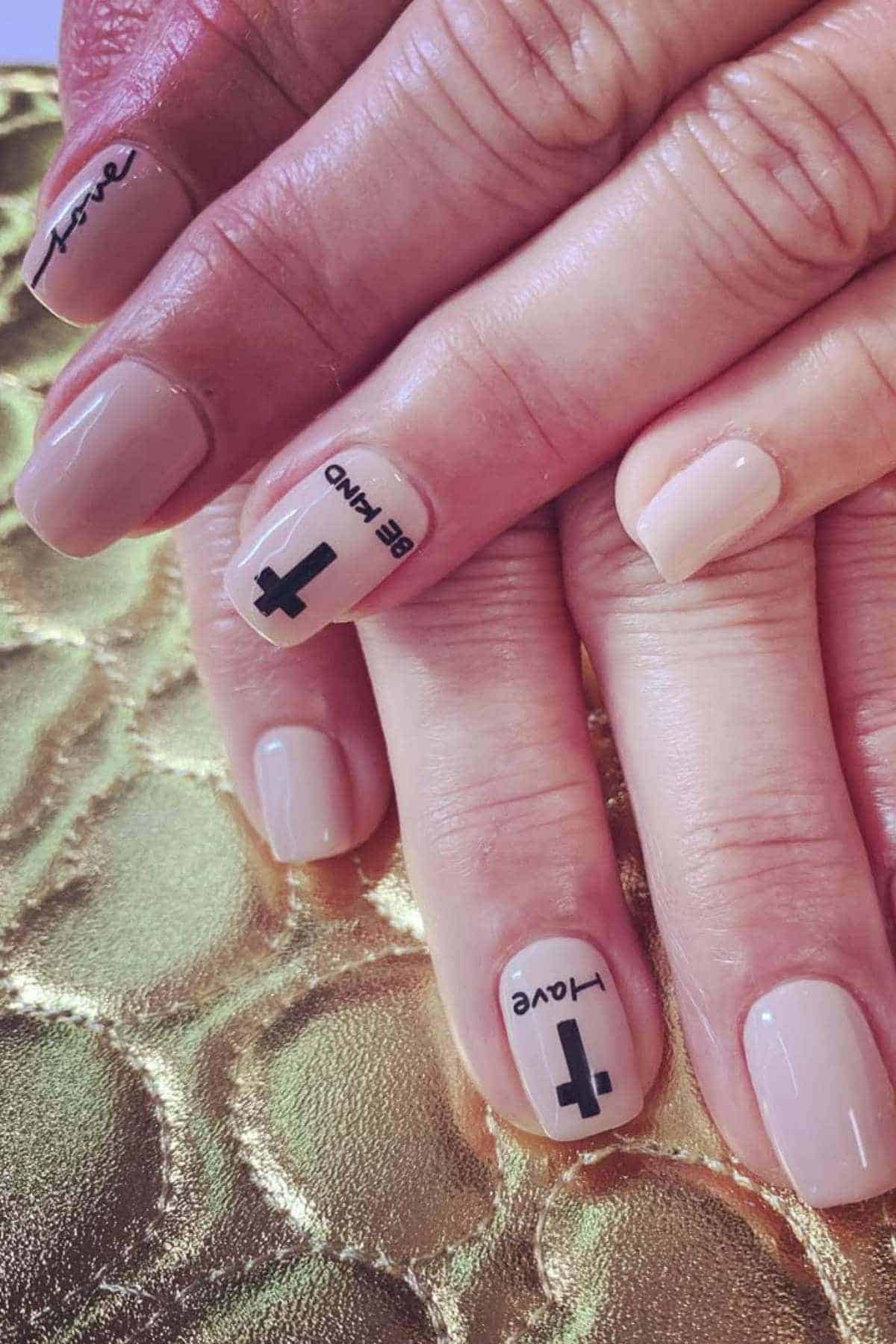 Two hands with nude manicure; index nails have black crosses and words “BE KIND” and “love” printed in black. Nails rest on a textured gold surface.