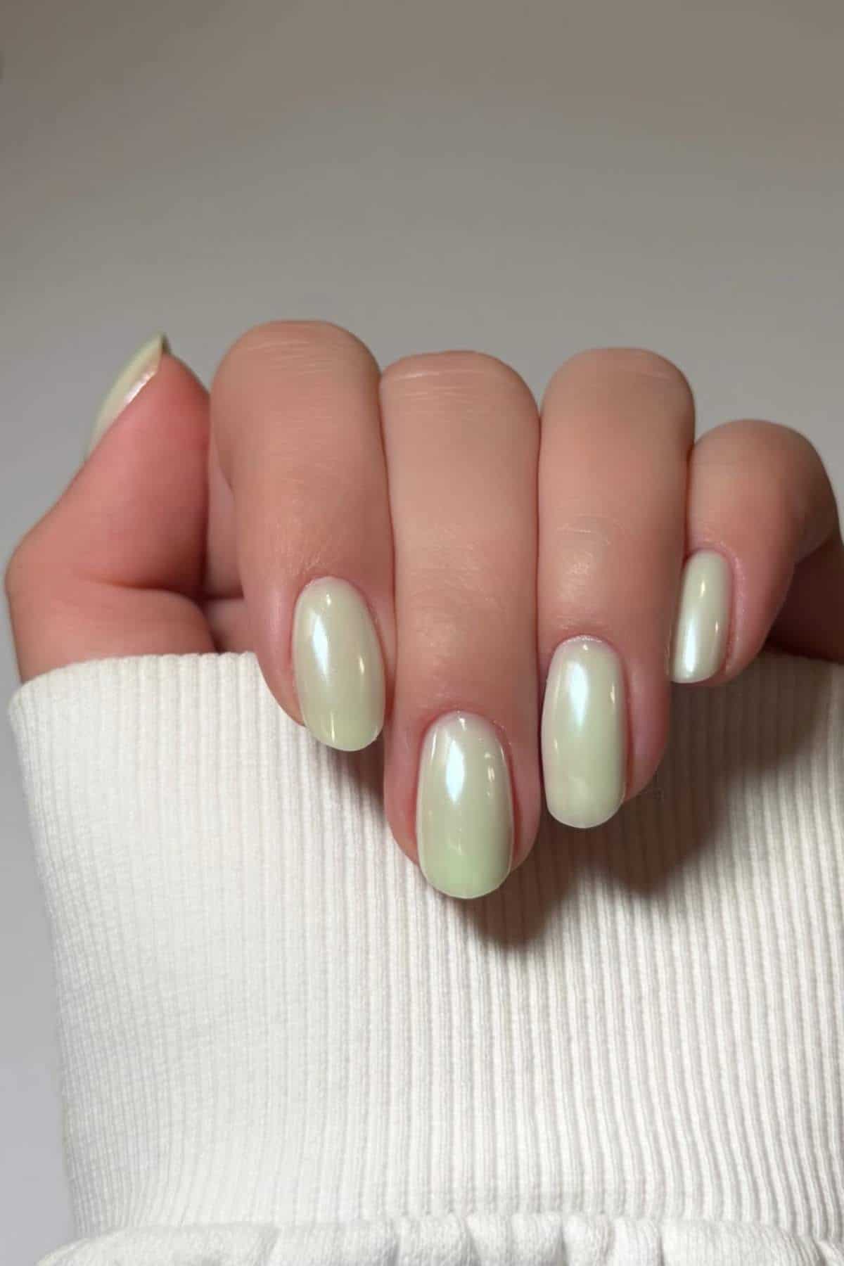 A hand with short, oval-shaped nails painted in a pale, glossy pastel green shade, partially covered by a white ribbed sleeve.