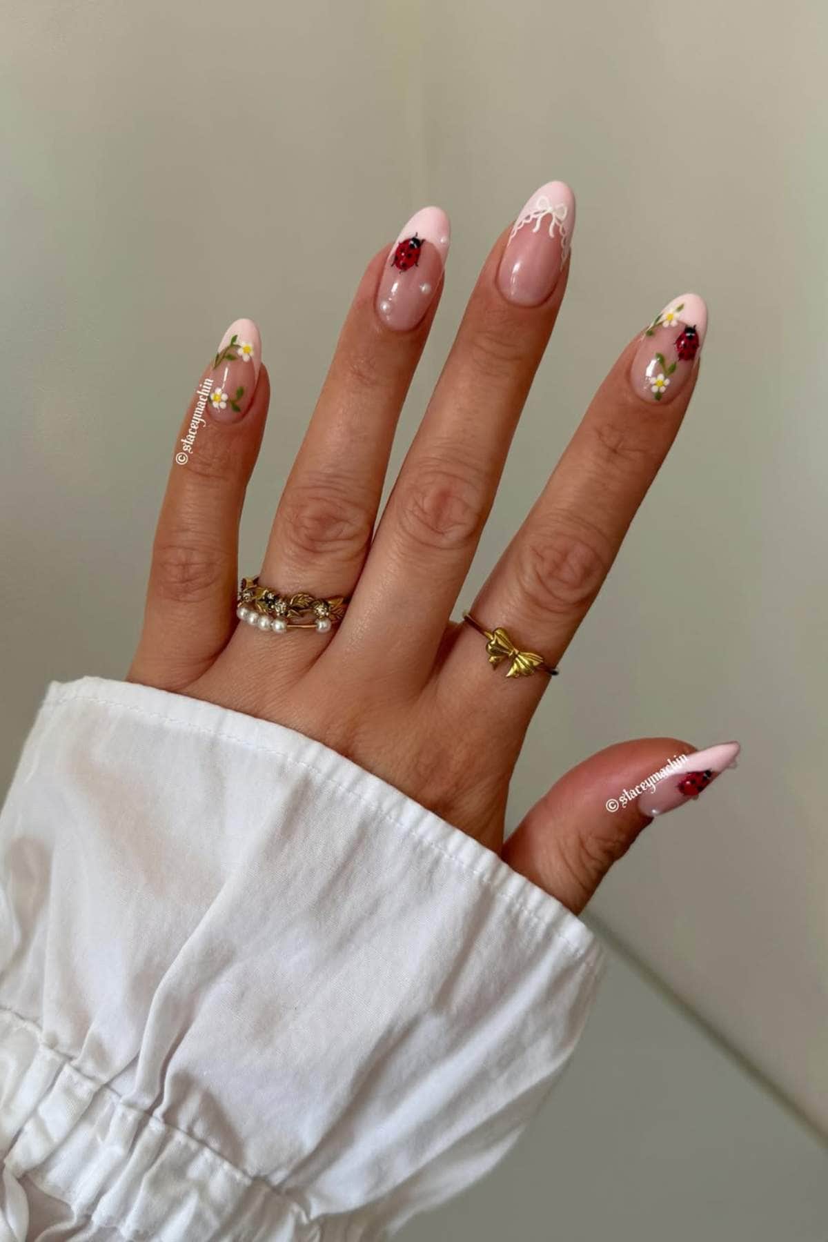 A hand with pink, almond-shaped nails featuring floral and lace designs, wearing three gold rings, shown against a plain background.