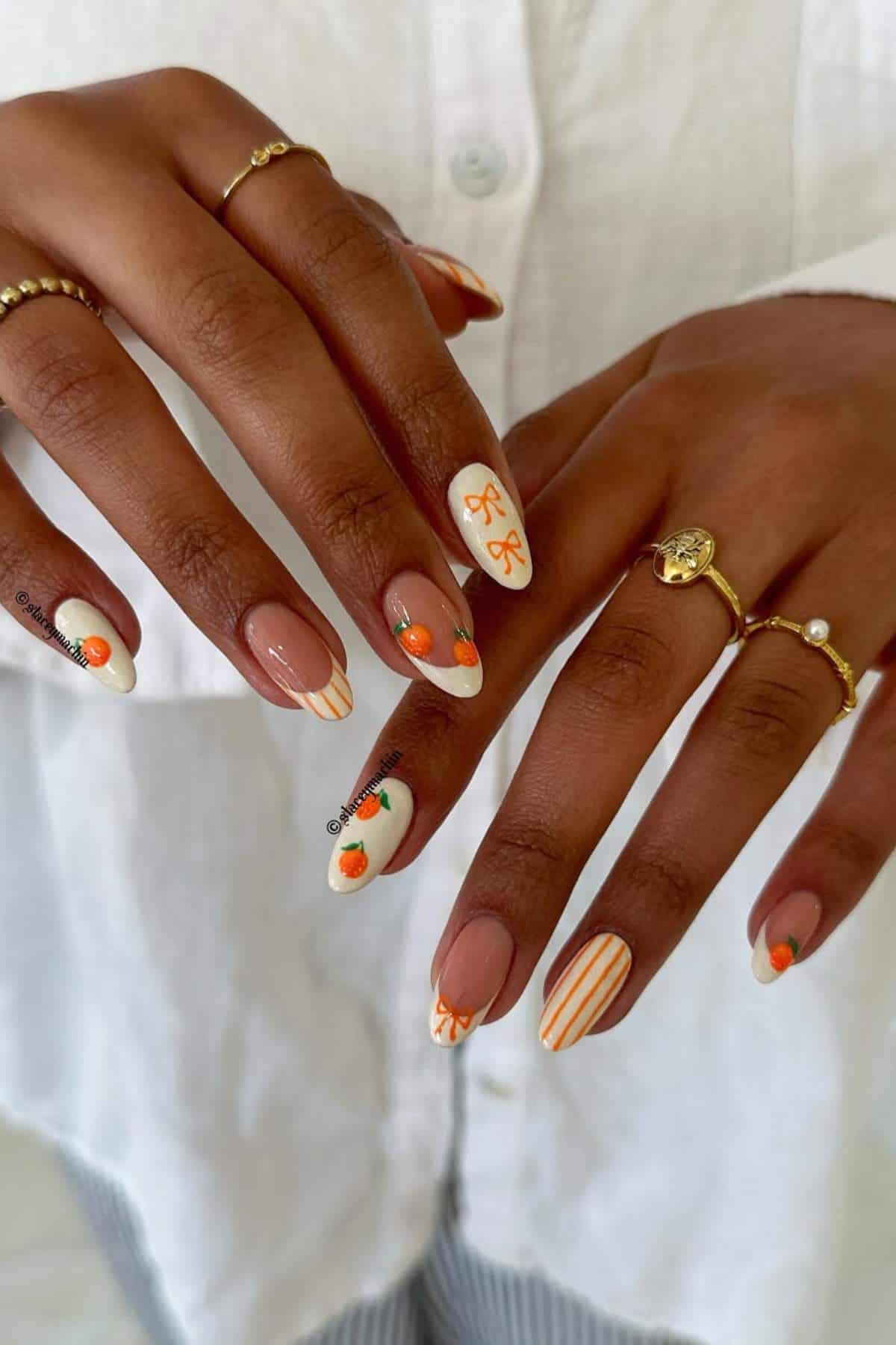 A person with neatly manicured nails featuring orange and white designs, including stripes, flowers, and abstract patterns, wearing gold rings on several fingers.