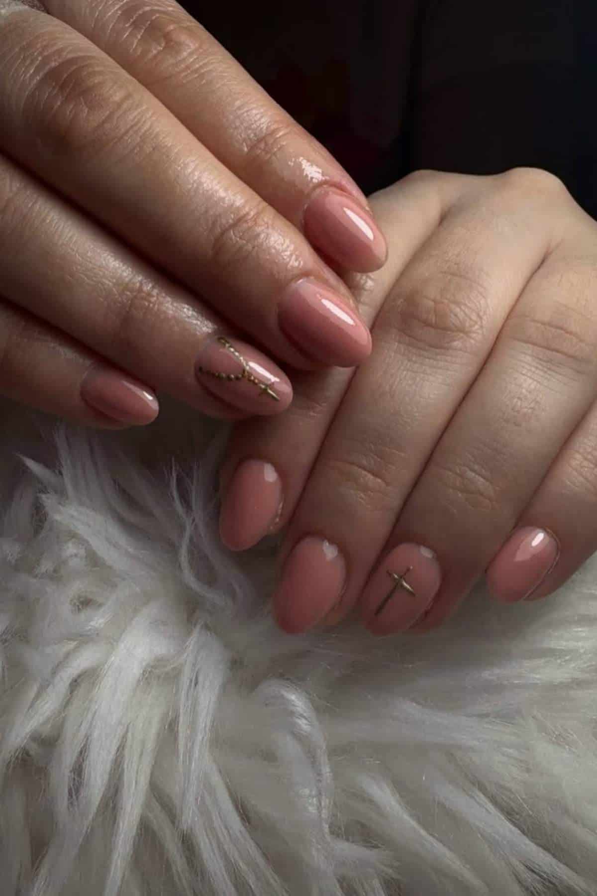 Hands with short, almond-shaped nude pink nails featuring minimal gold line and cross designs on the ring fingernails, resting on a white fluffy surface.