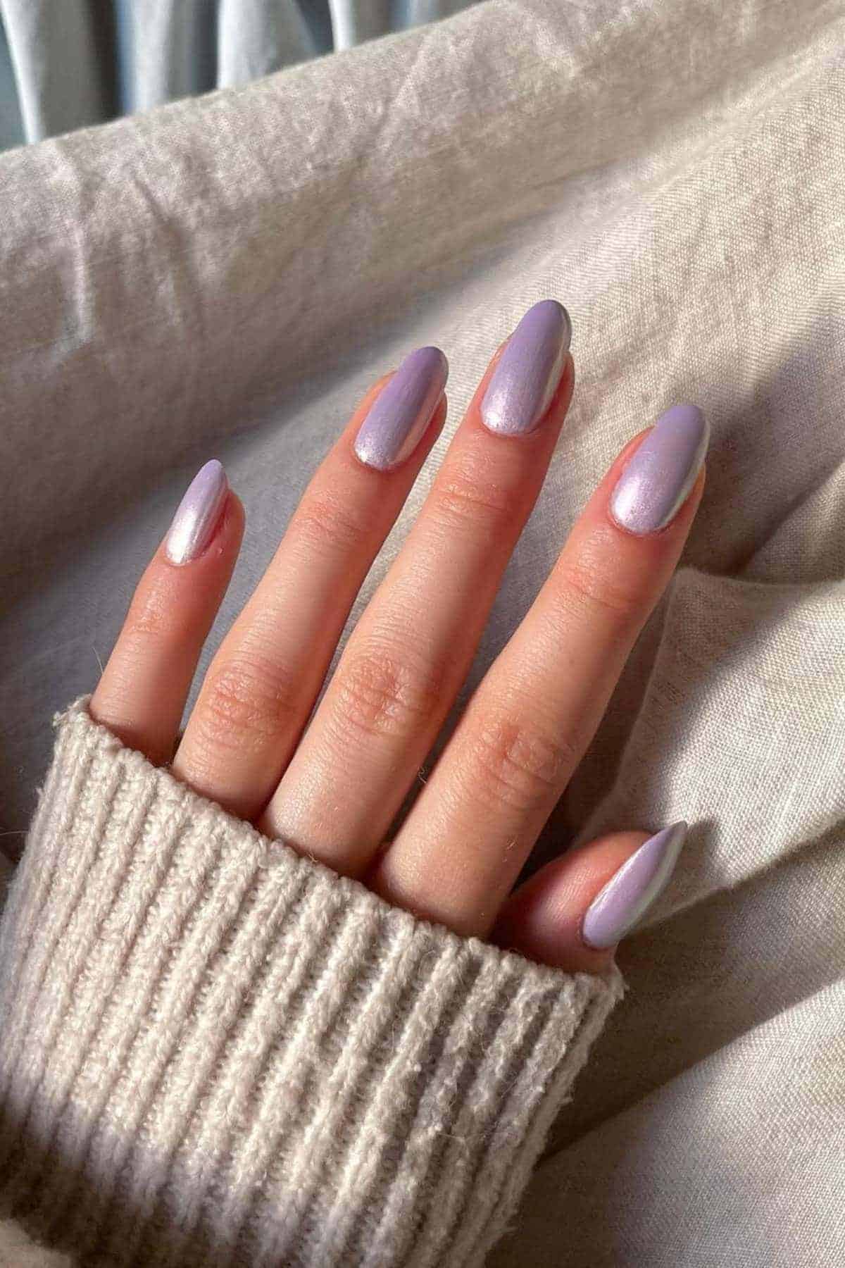 A hand with neatly manicured nails painted in a pastel lavender color rests on a light beige fabric, with a beige knitted sleeve partially covering the wrist.