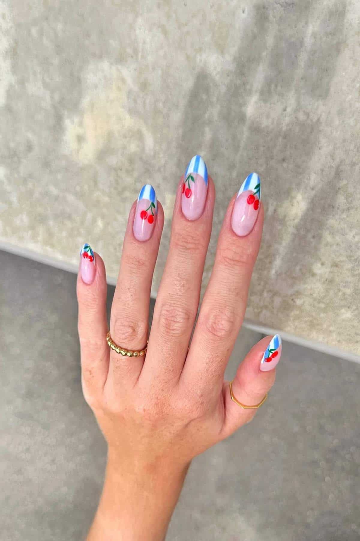 A hand with blue French tip nails featuring small red cherry designs, wearing two gold rings, resting against a light, textured background.