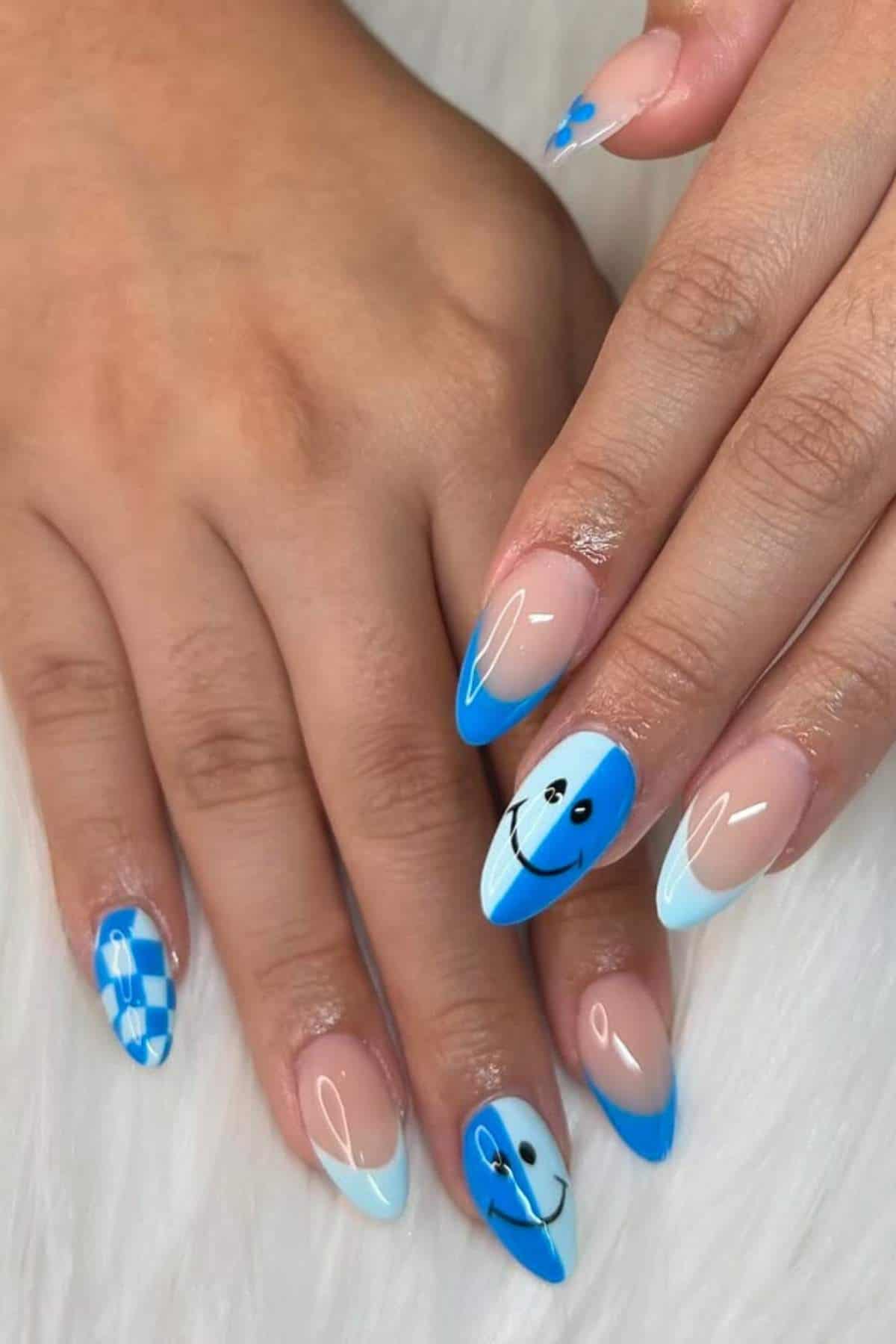 A hand with almond-shaped nails features blue and light blue nail art, including French tips, smiley faces, and a checkered pattern on a white background.