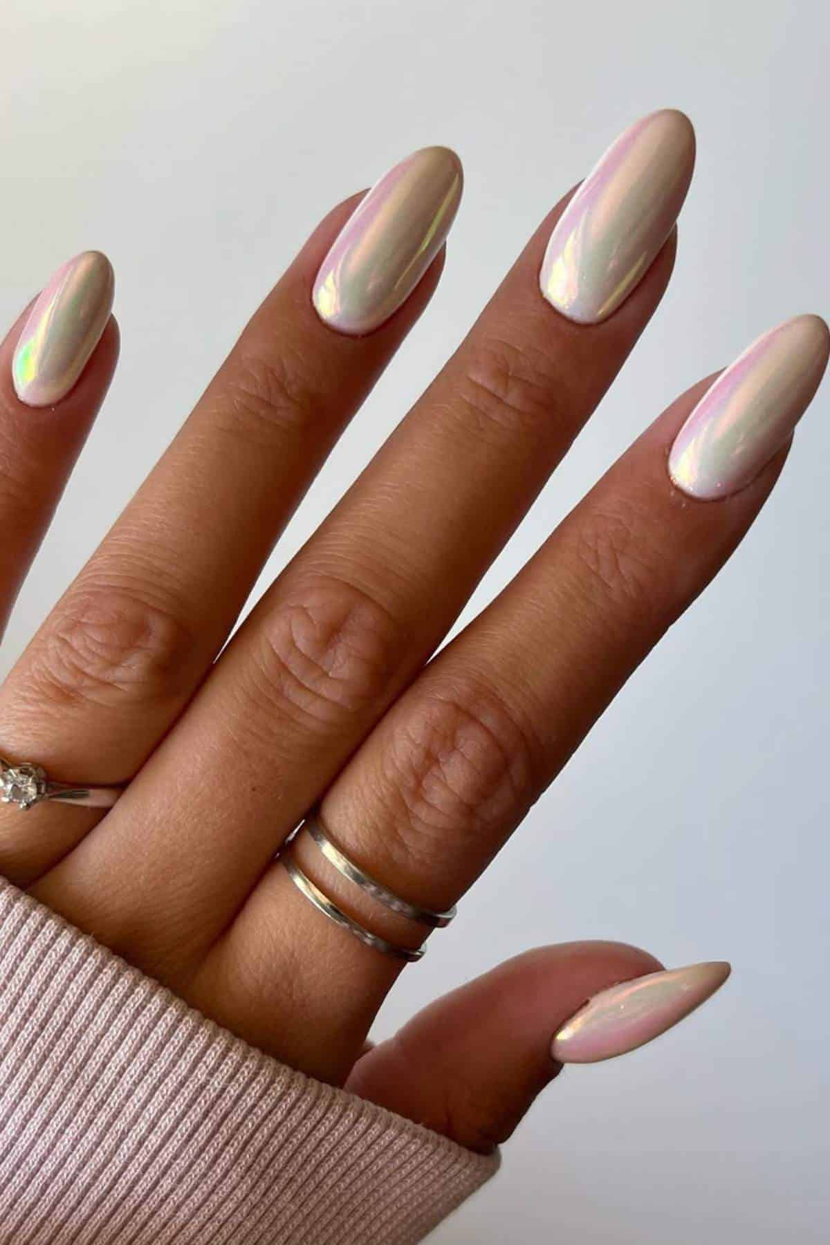 A hand with long, almond-shaped nails painted with a pearlescent, iridescent polish, wearing two rings on the ring finger and a light pink sweater sleeve partially visible.
