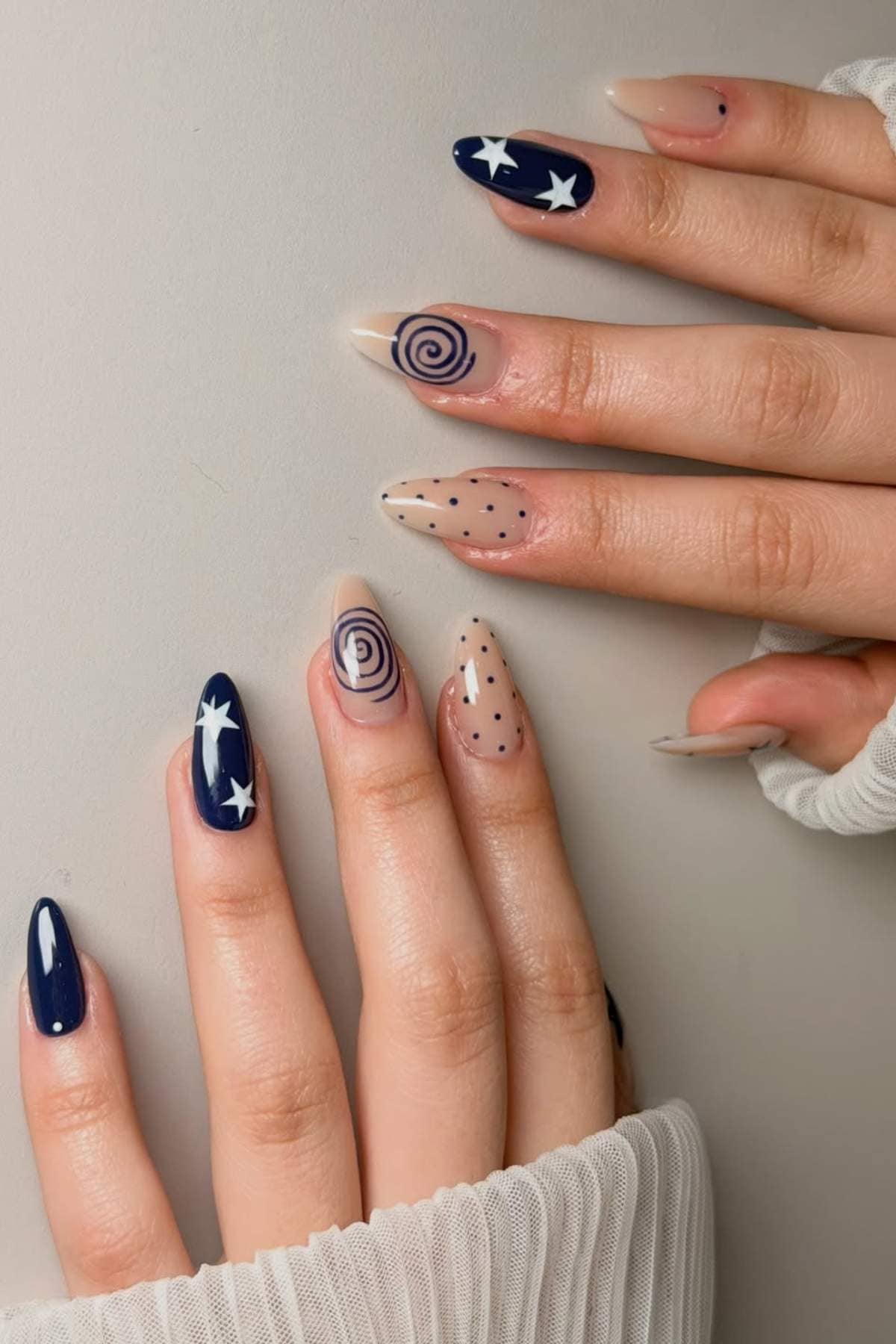 A hand with almond-shaped nails features navy blue, white stars, black dots, and spiral designs on a mix of nude and dark blue backgrounds.