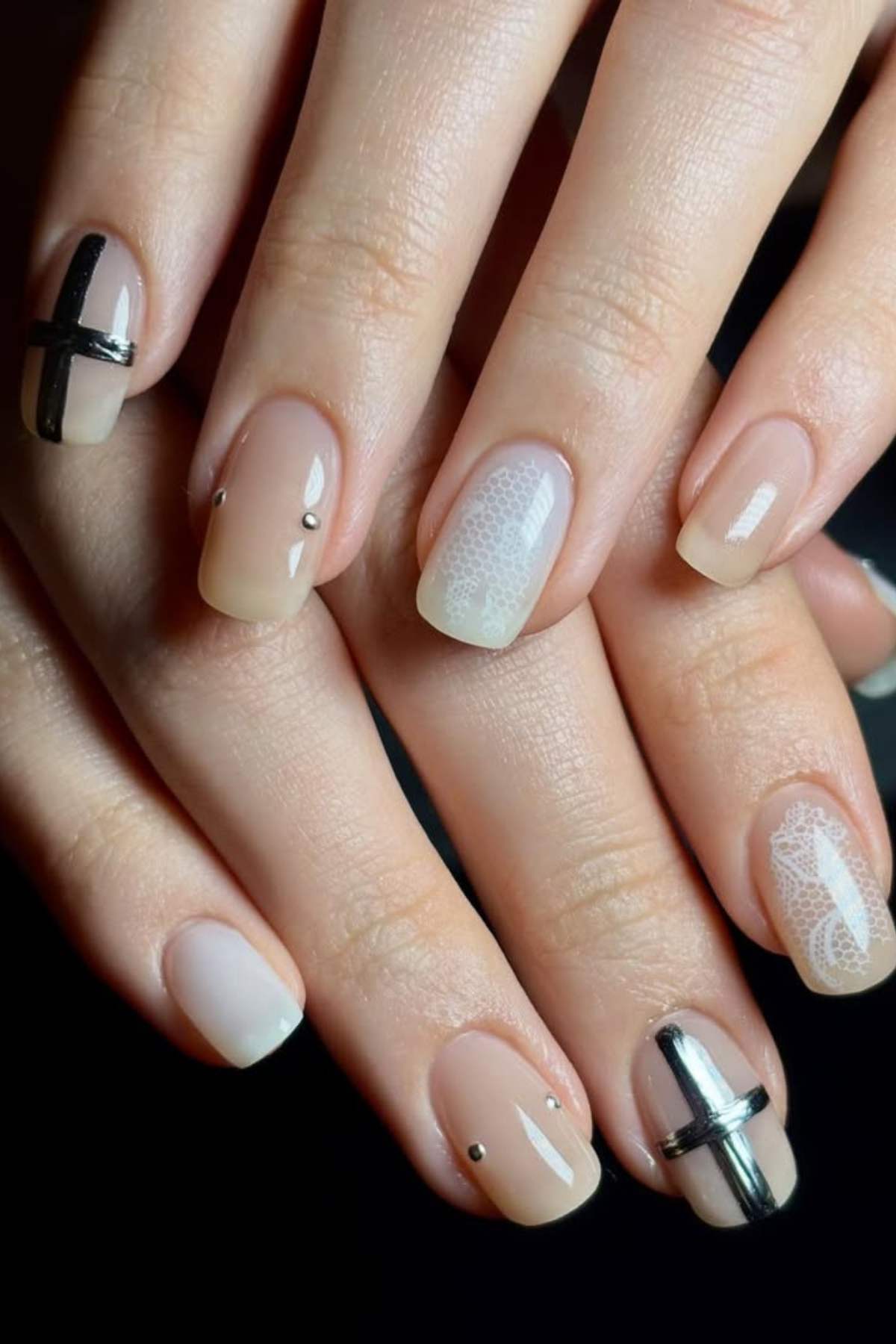 A close-up of hands with nude manicure featuring minimal black and silver line designs, small silver studs, and white lace-like patterns on some nails.