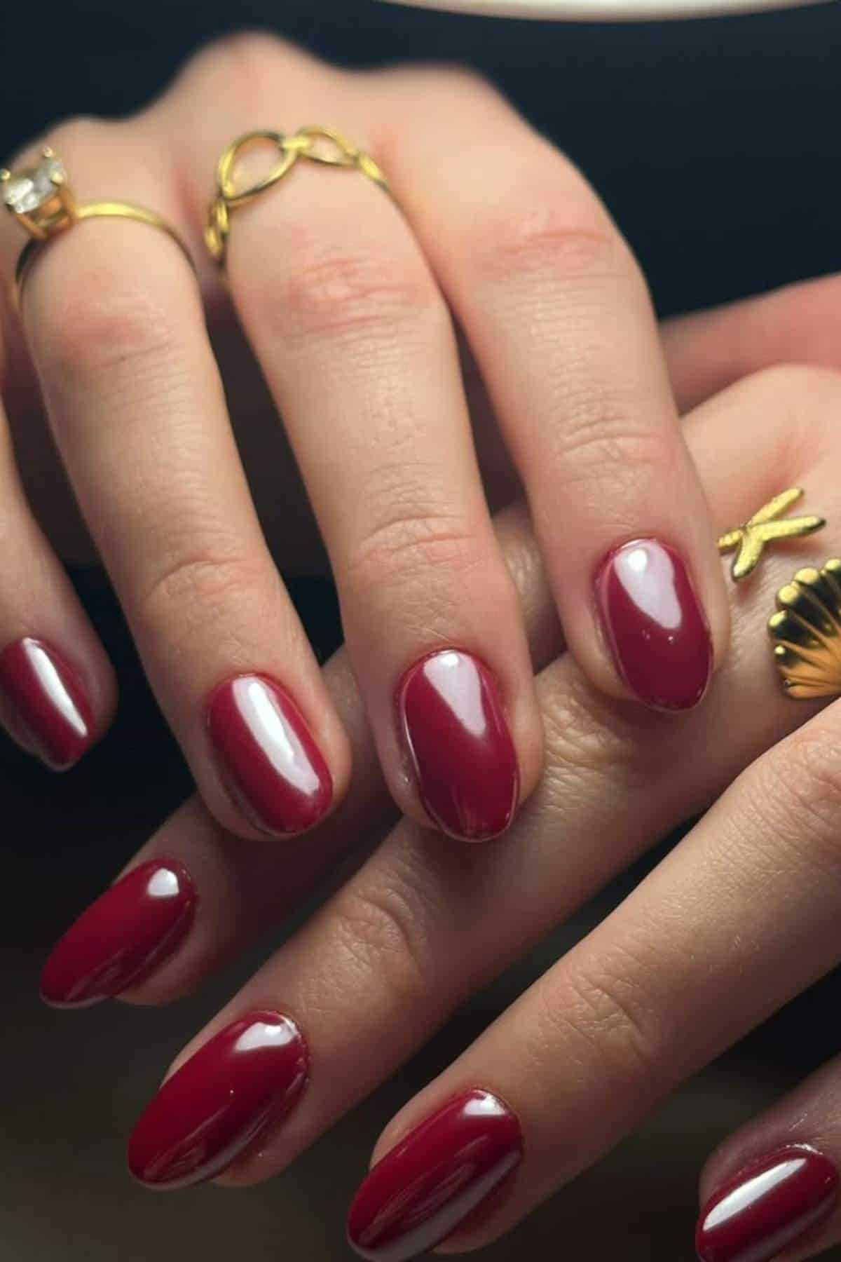 Close-up of hands with glossy red nail polish and gold rings, including one shaped like a shell and another with an infinity symbol.