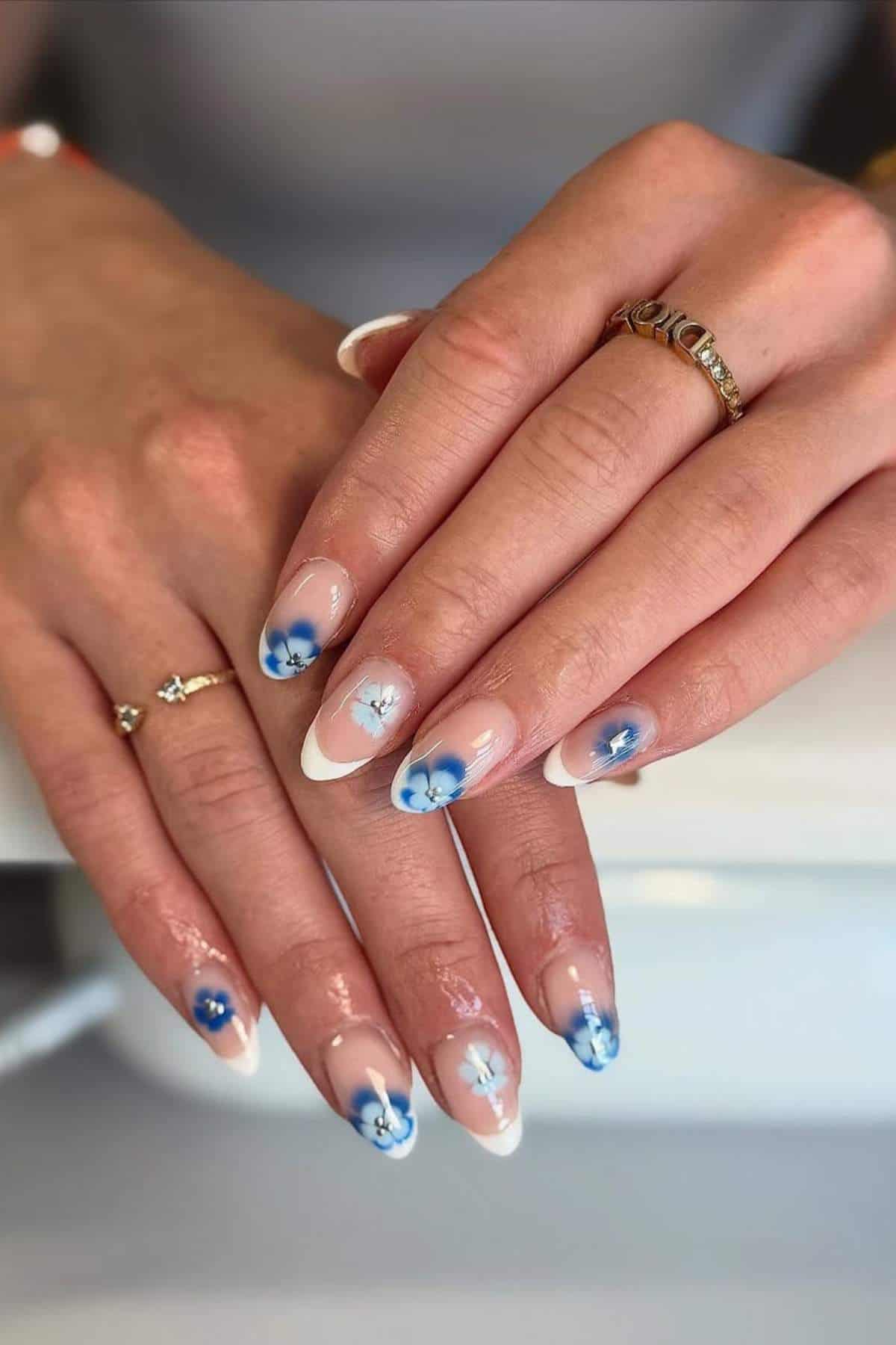 Hands with almond-shaped nails featuring a French tip design and blue and white floral nail art. The person is wearing gold rings on their fingers.