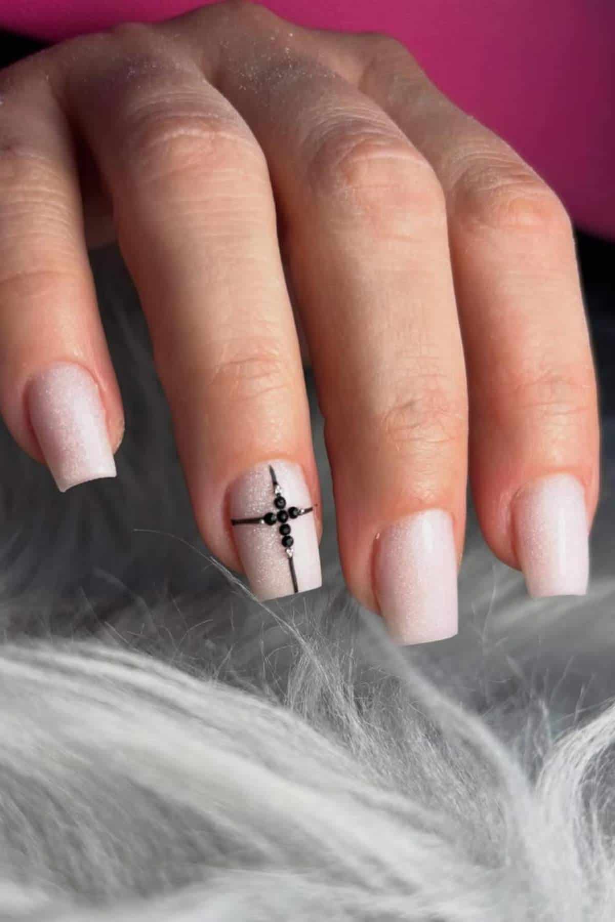 A hand with short, square-shaped light pink nails; the ring finger features a black cross design with small black dots.