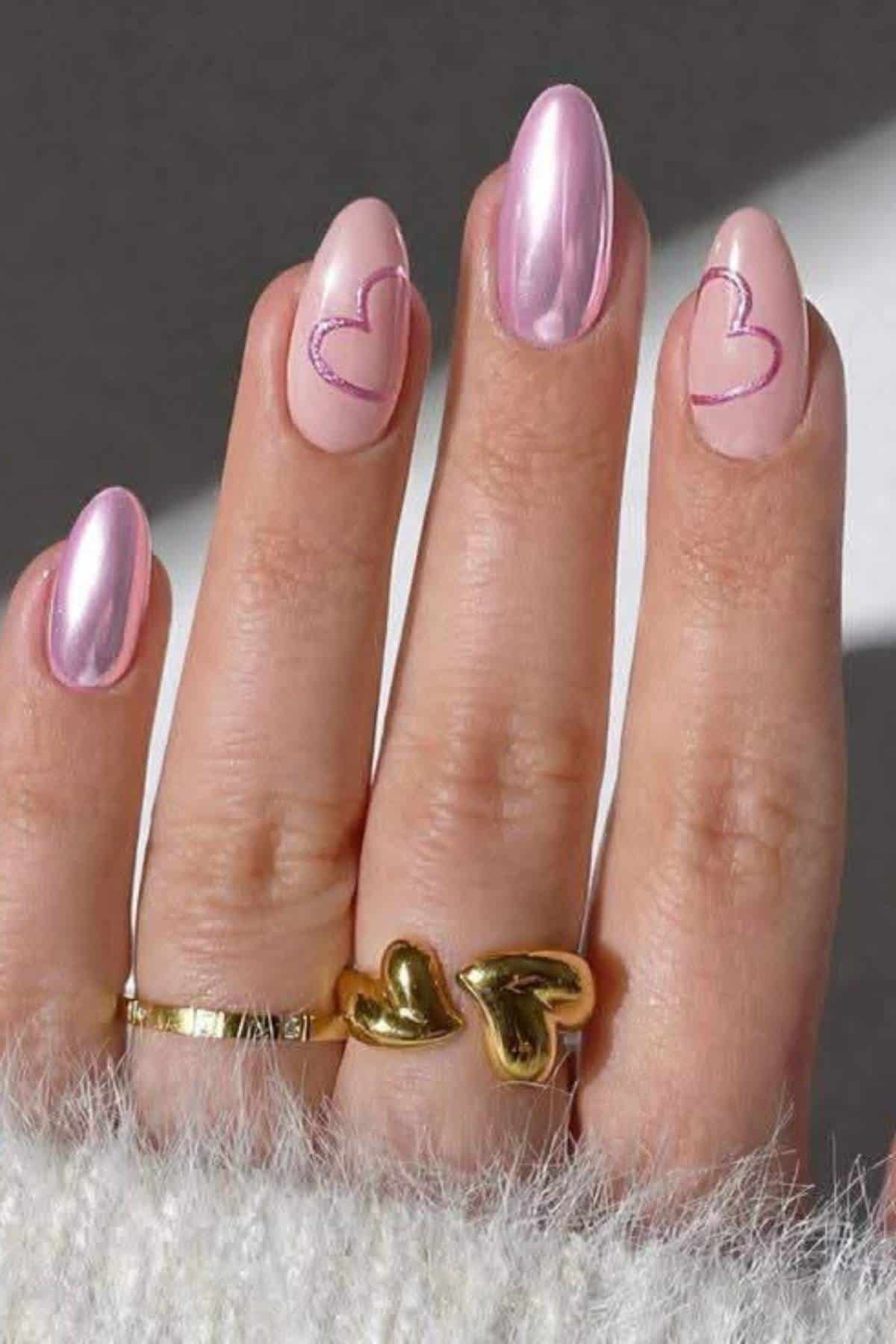 A hand with long, almond-shaped pink nails, two featuring heart designs. The person wears gold rings, one with a heart shape, and a white fluffy sleeve is partially visible.