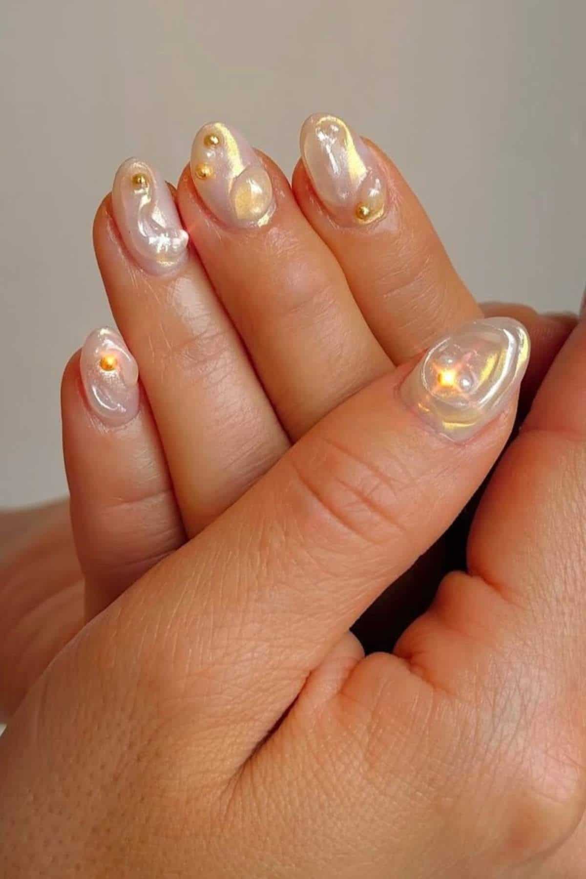 Close-up of hands with iridescent, pearl-like nail art featuring raised gel designs and small gold embellishments on each nail.