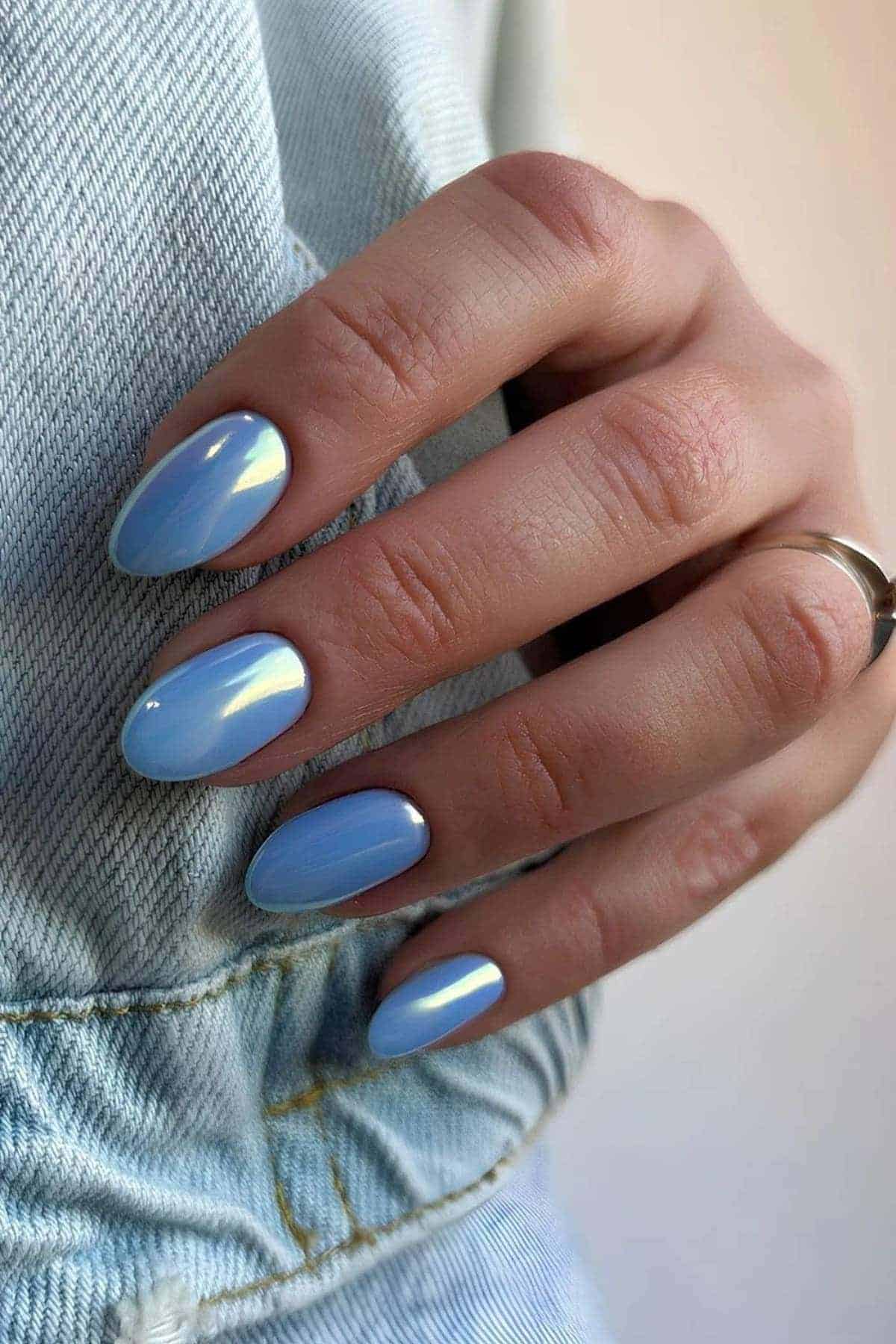 A hand with almond-shaped nails painted in glossy light blue, resting on light denim fabric. The person is wearing a simple silver ring.