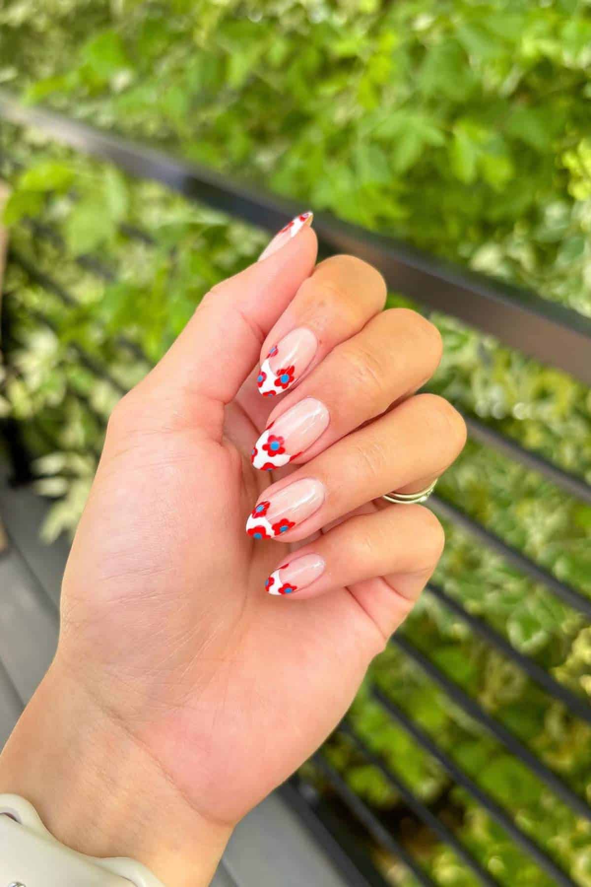 A hand with almond-shaped nails features a nude base and red flower designs near the tips, held against a background of green foliage and a black railing.