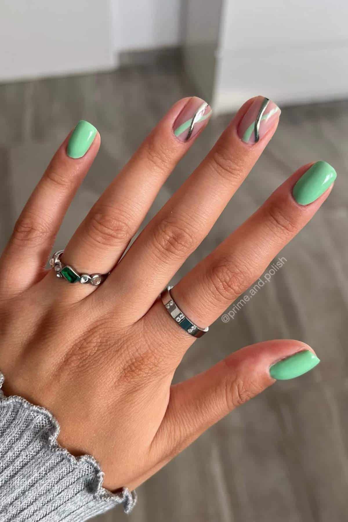 A hand with mint green nail polish; accent nails feature abstract wavy designs in green, white, and beige. The person wears two rings and a ribbed sleeve.