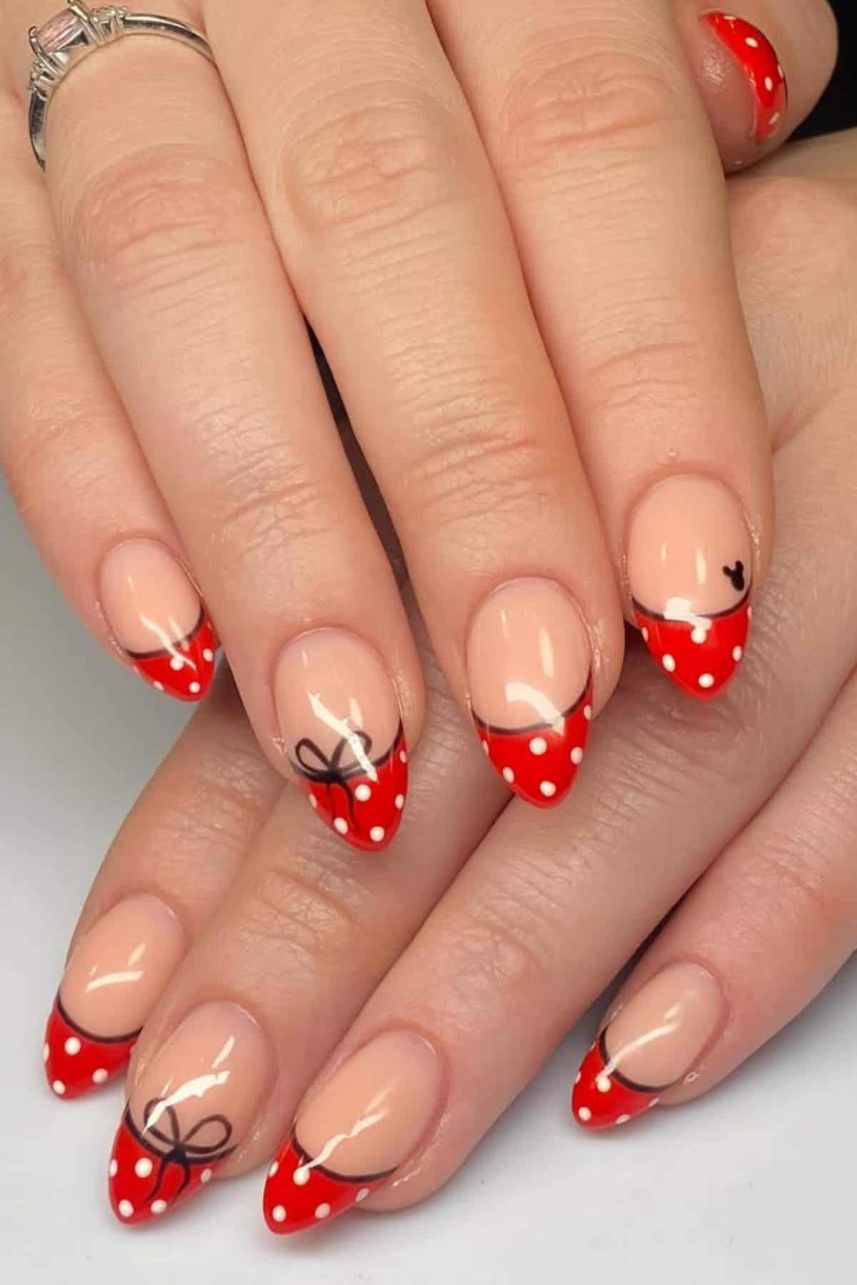 Hands with almond-shaped nails featuring red French tips with white polka dots, black bow designs, and a small black heart accent on a natural pink base.
