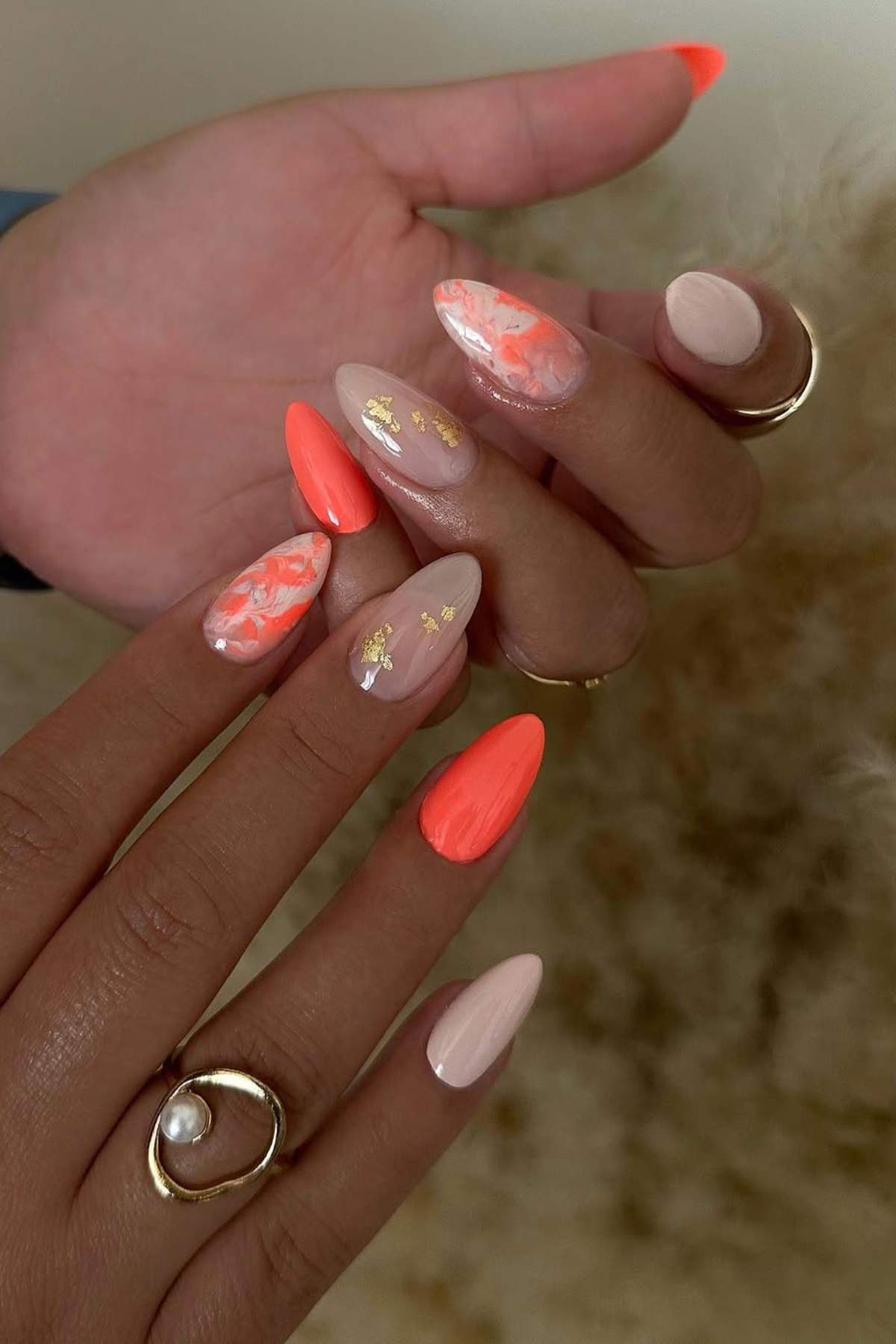 A hand with long almond-shaped nails painted in a mix of nude, orange, marble, and gold foil designs, holding another hand with rings.