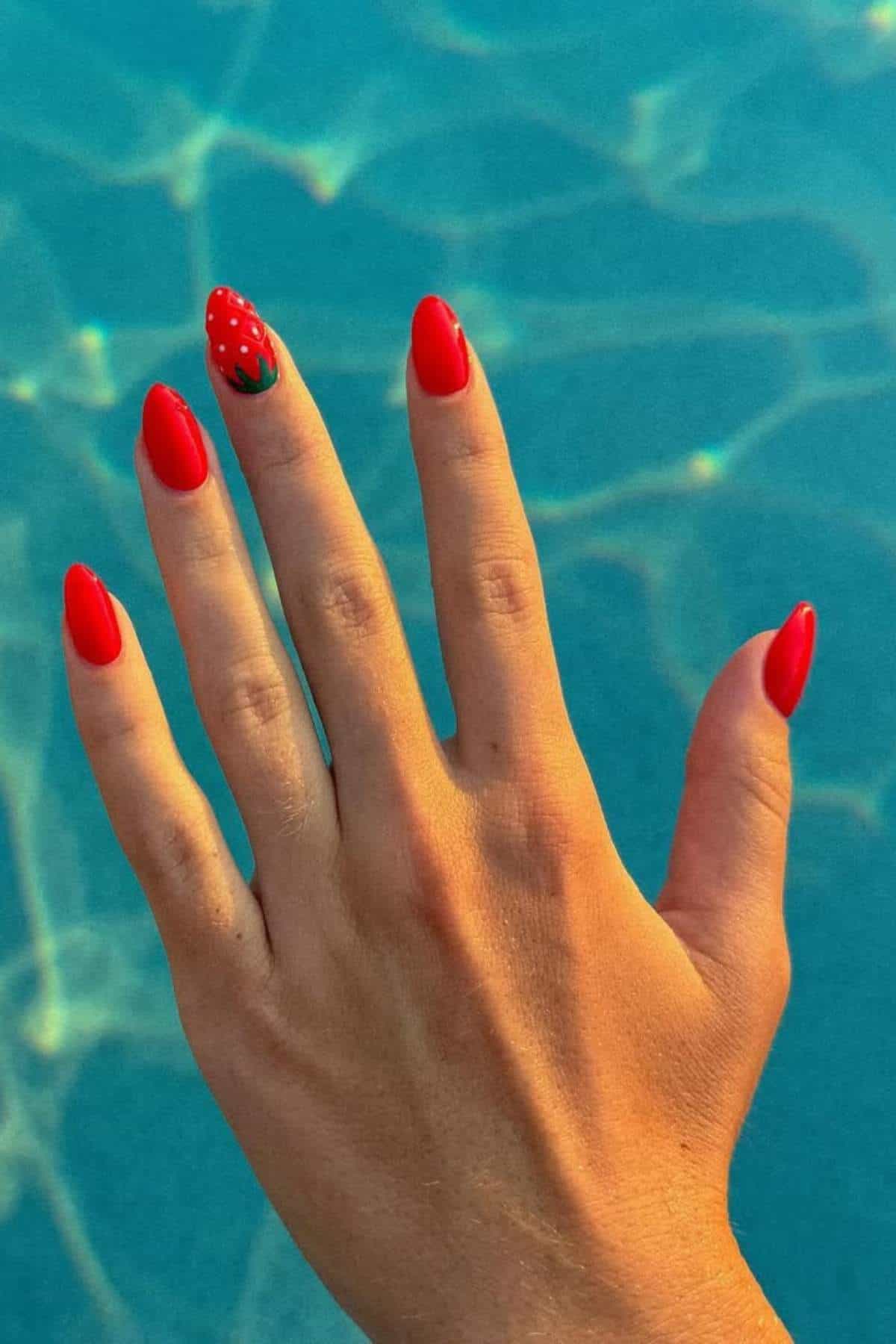 A hand with red, almond-shaped nails, one featuring a strawberry design, is held above a blue, sunlit pool.
