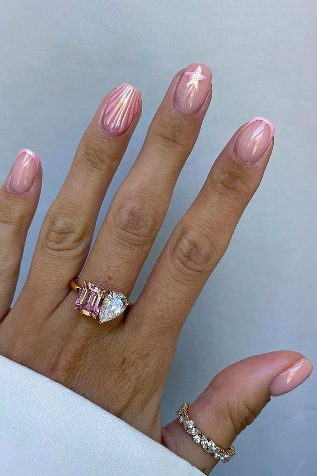 A hand with pink and white nail art, wearing gold rings with pink and clear gemstones, against a light background.