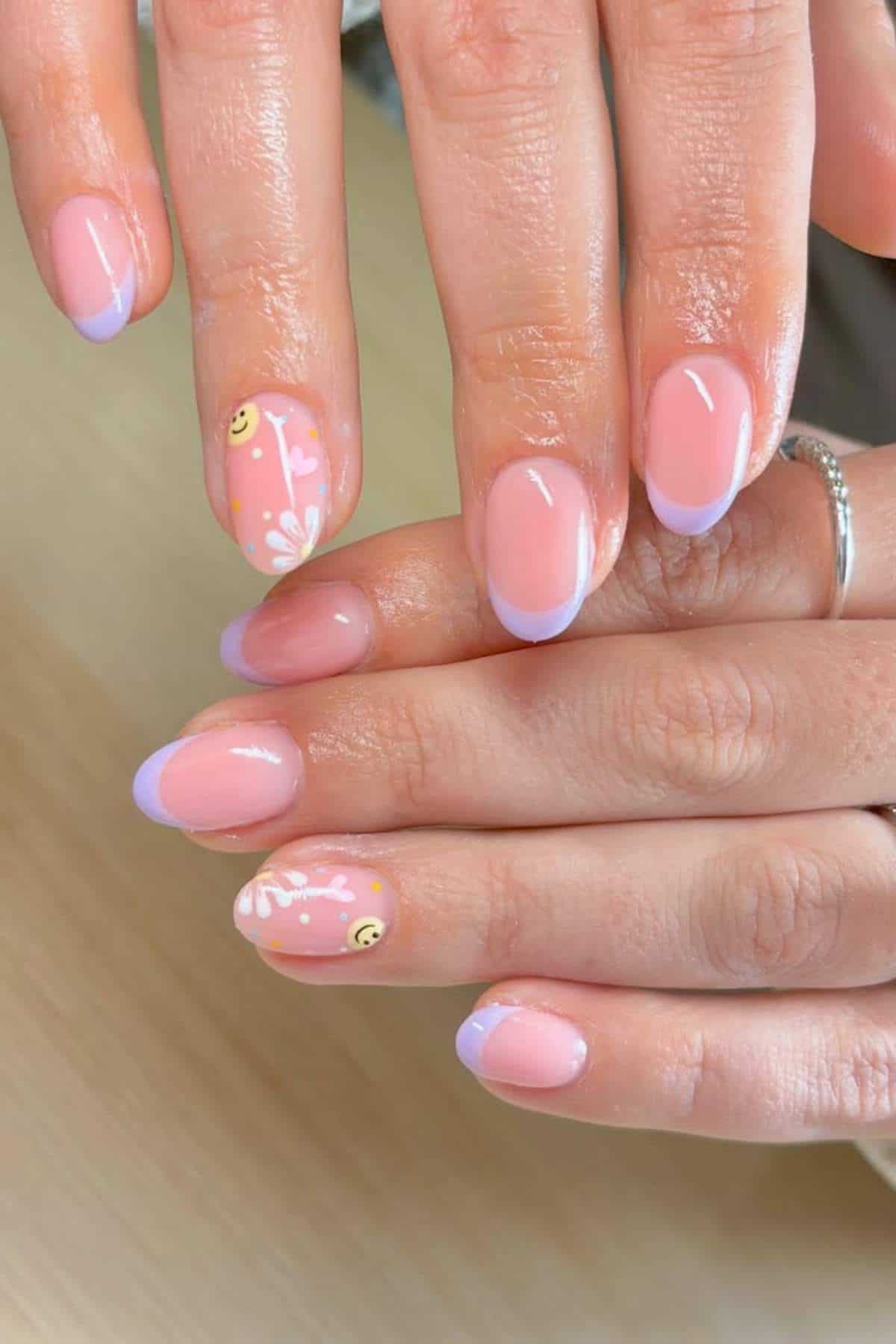 Hands with almond-shaped nails featuring a light pink base, pale purple French tips, and accent nails with white floral designs and small yellow smiley faces.