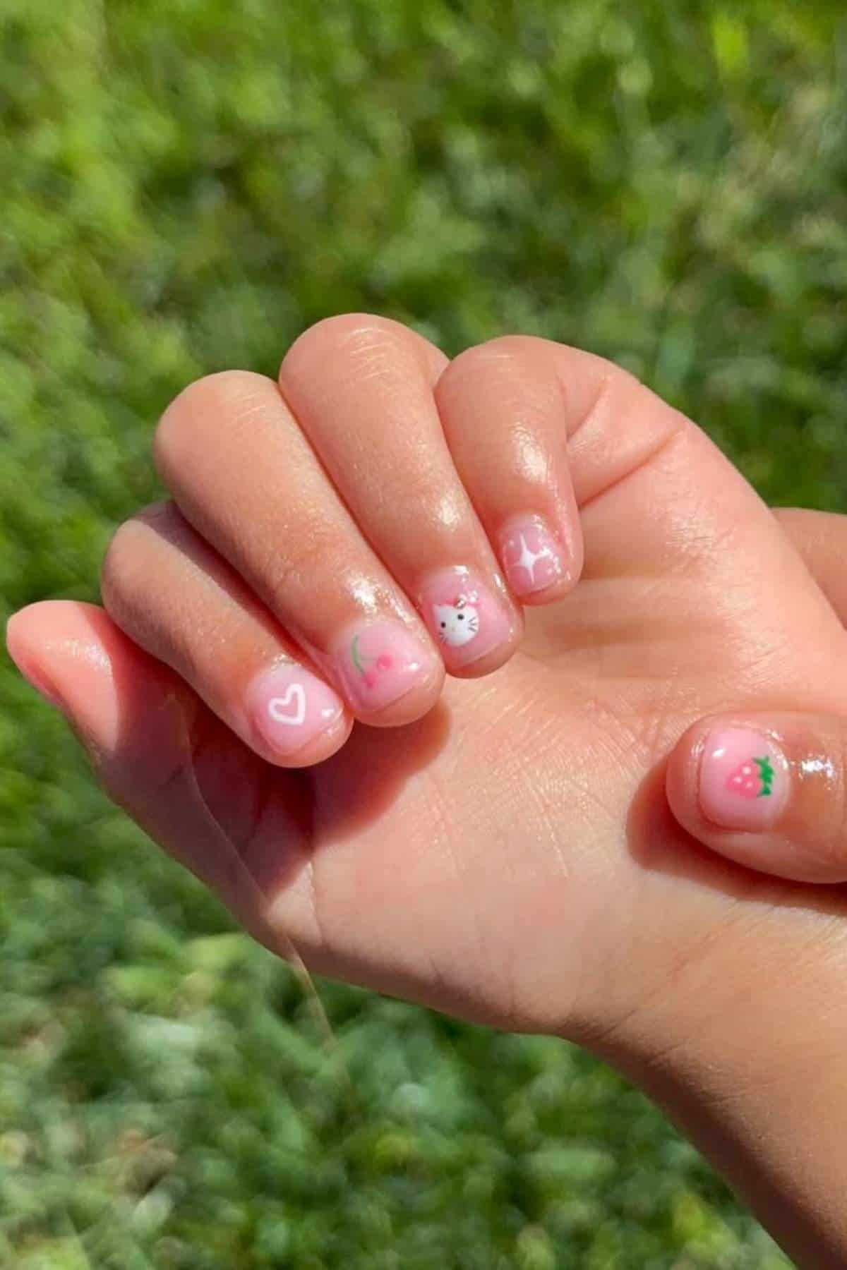 A hand with short nails painted with pink polish and decorated with small designs, including a heart, a cat face, and strawberries, held against a grassy background.
