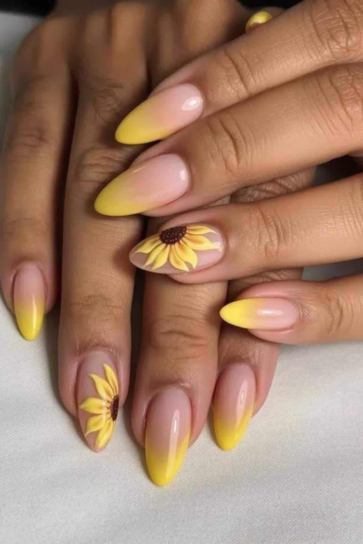 Hands with almond-shaped nails featuring yellow ombre tips and sunflower designs on two accent nails, resting on a white surface.