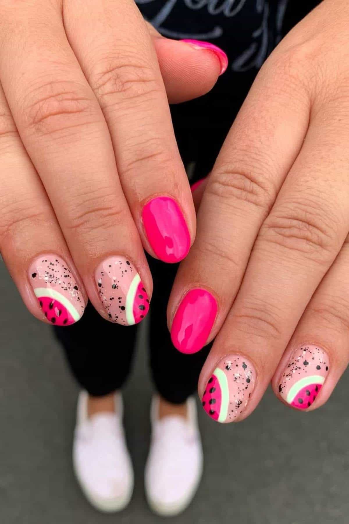 Hands with manicured nails featuring bright pink polish and accent nails with watermelon-inspired designs and black glitter. White shoes and dark pants are visible in the background.