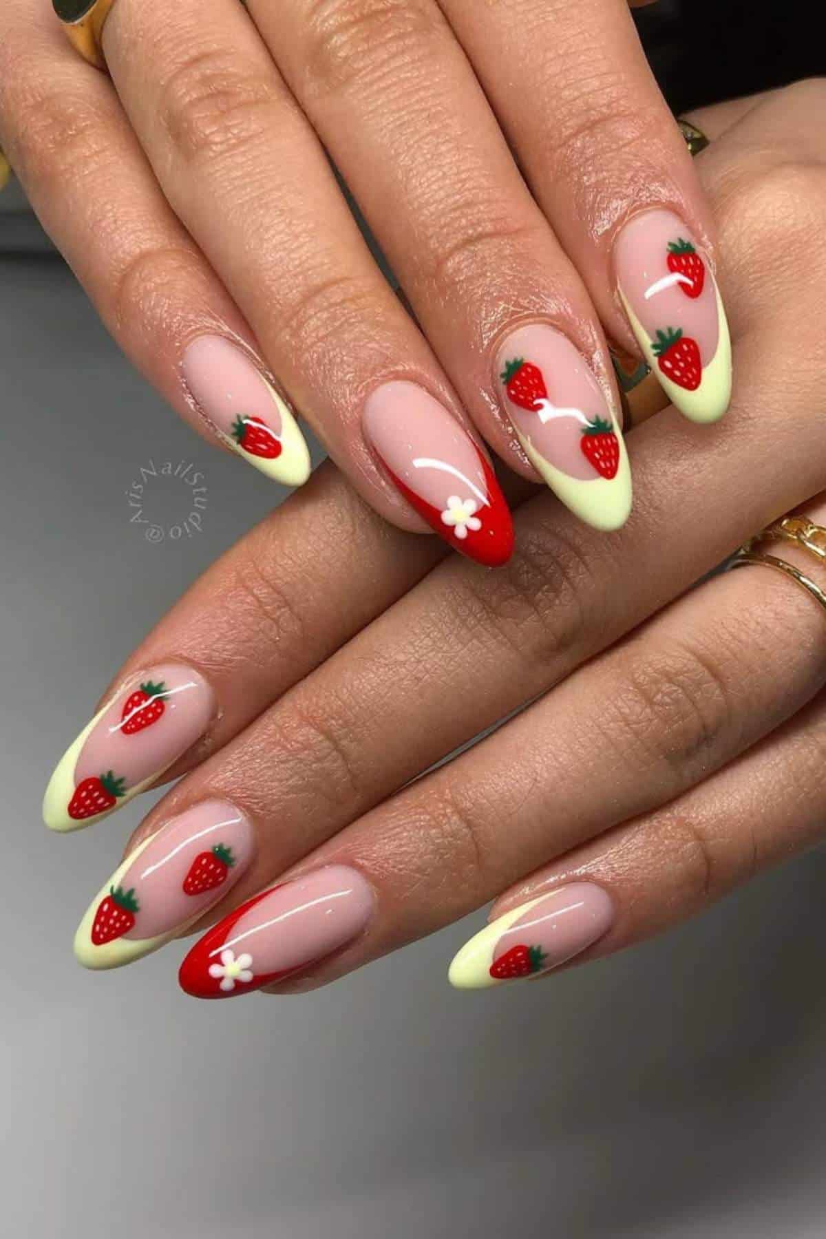 Hands with almond-shaped nails featuring a light yellow tip, red strawberries, green leaves, and a white flower design on a nude pink base.