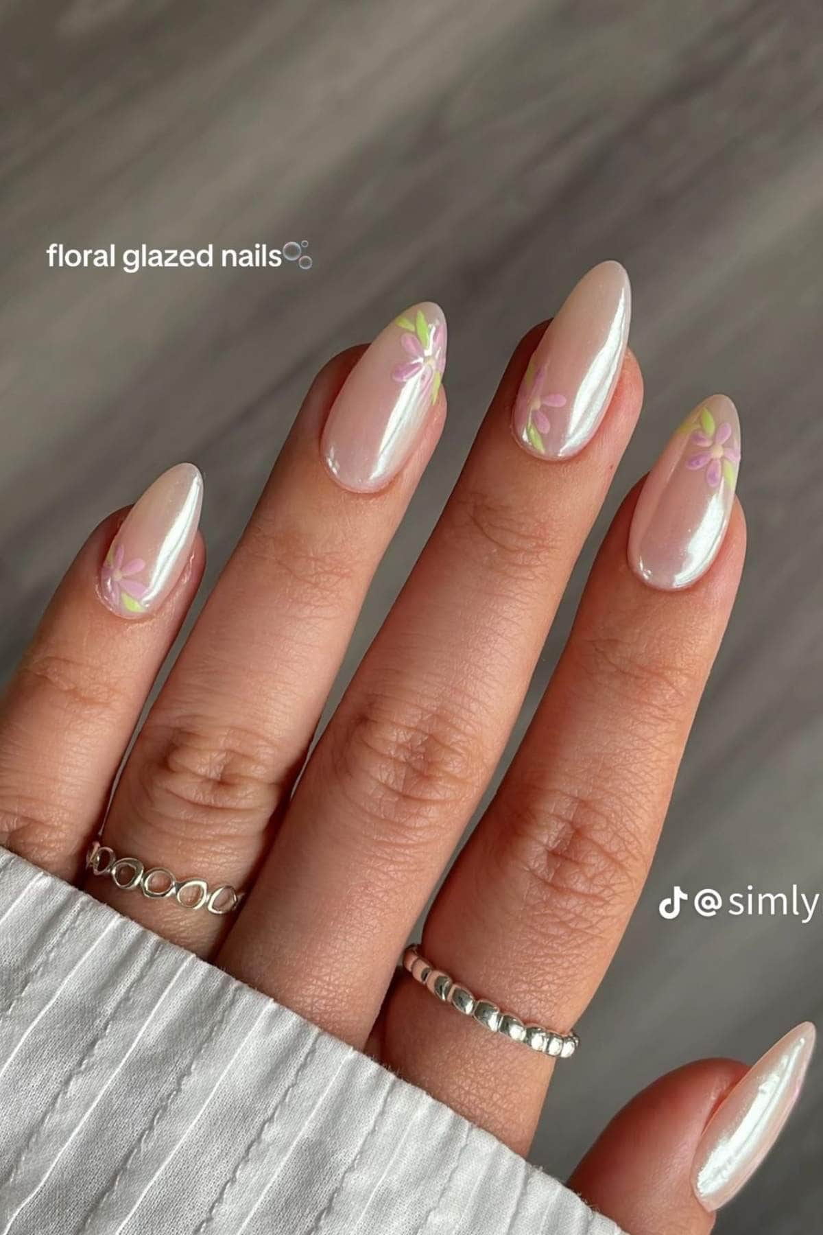 A hand with almond-shaped nails features a pearly glazed finish and small pastel flower designs near the tips. The person is wearing silver rings.