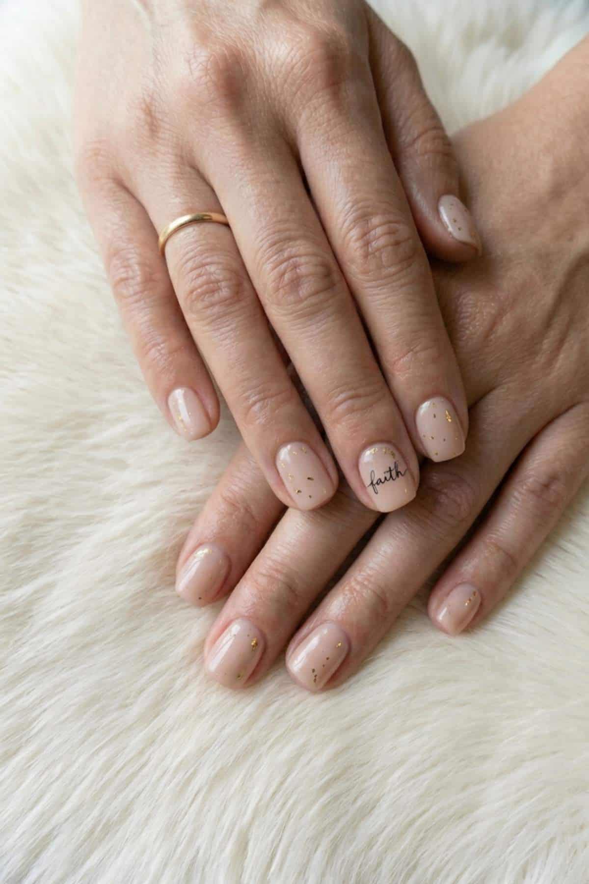 Two hands with short, nude-colored polished nails, gold flakes on most nails, and the word "faith" written on one nail, resting on a white furry surface.