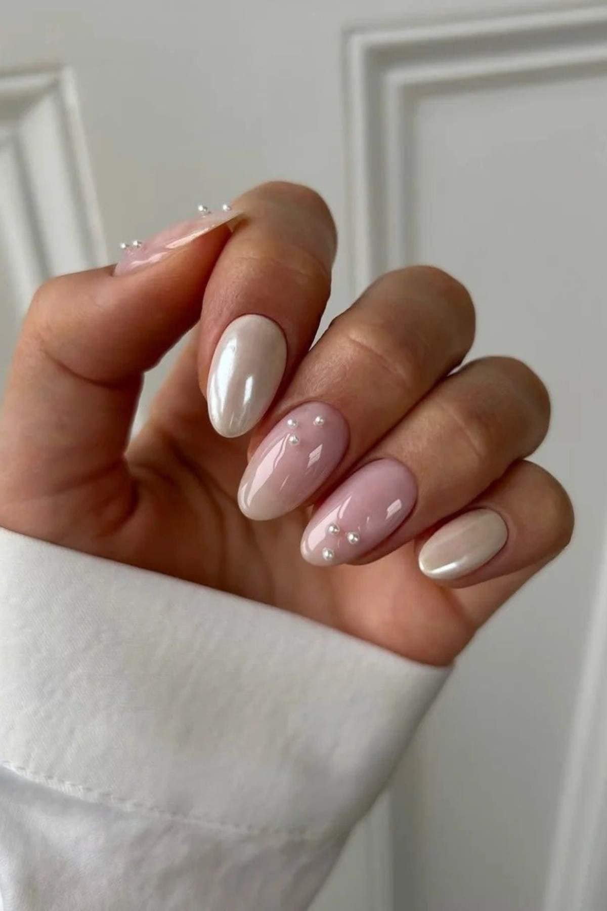 A hand with almond-shaped nails painted in pale pink and white, featuring glossy finish and small pearl decorations on two nails.
