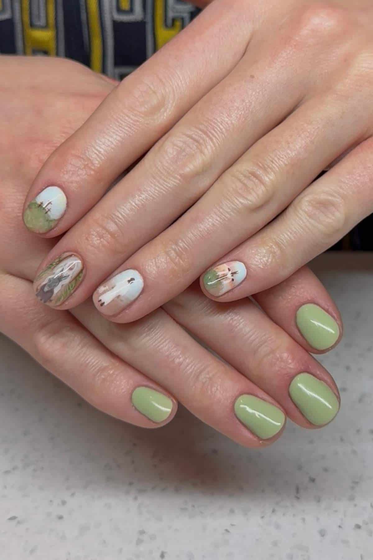 Hands with short, light green painted nails; two nails on each hand have a detailed landscape design featuring trees and a sky.