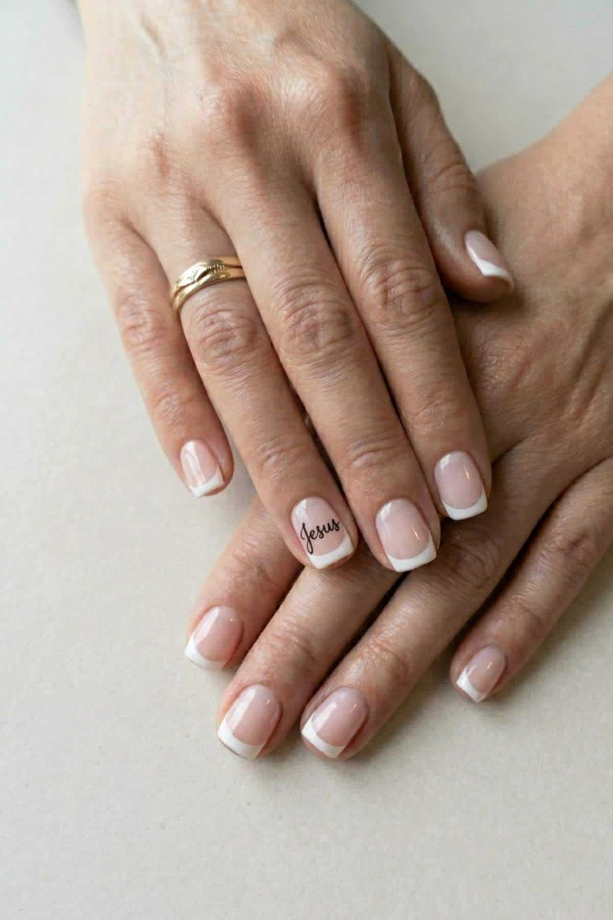 Close-up of hands with a French manicure; one ring on the left hand, and the word "Jesus" written on the ring fingernail.