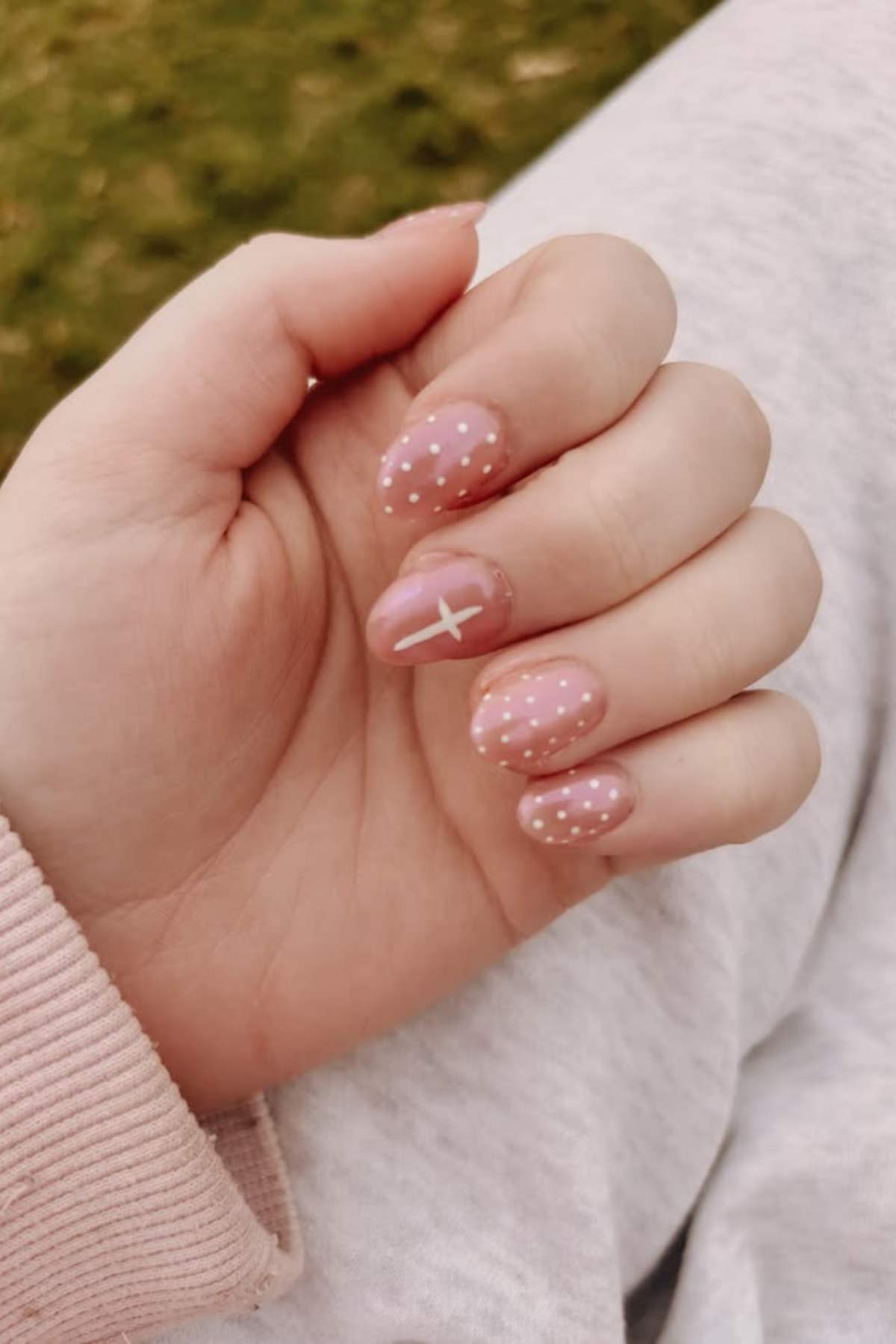 A hand with pink polished nails featuring white polka dots and a white cross design on the ring finger, resting on a pale fabric background.