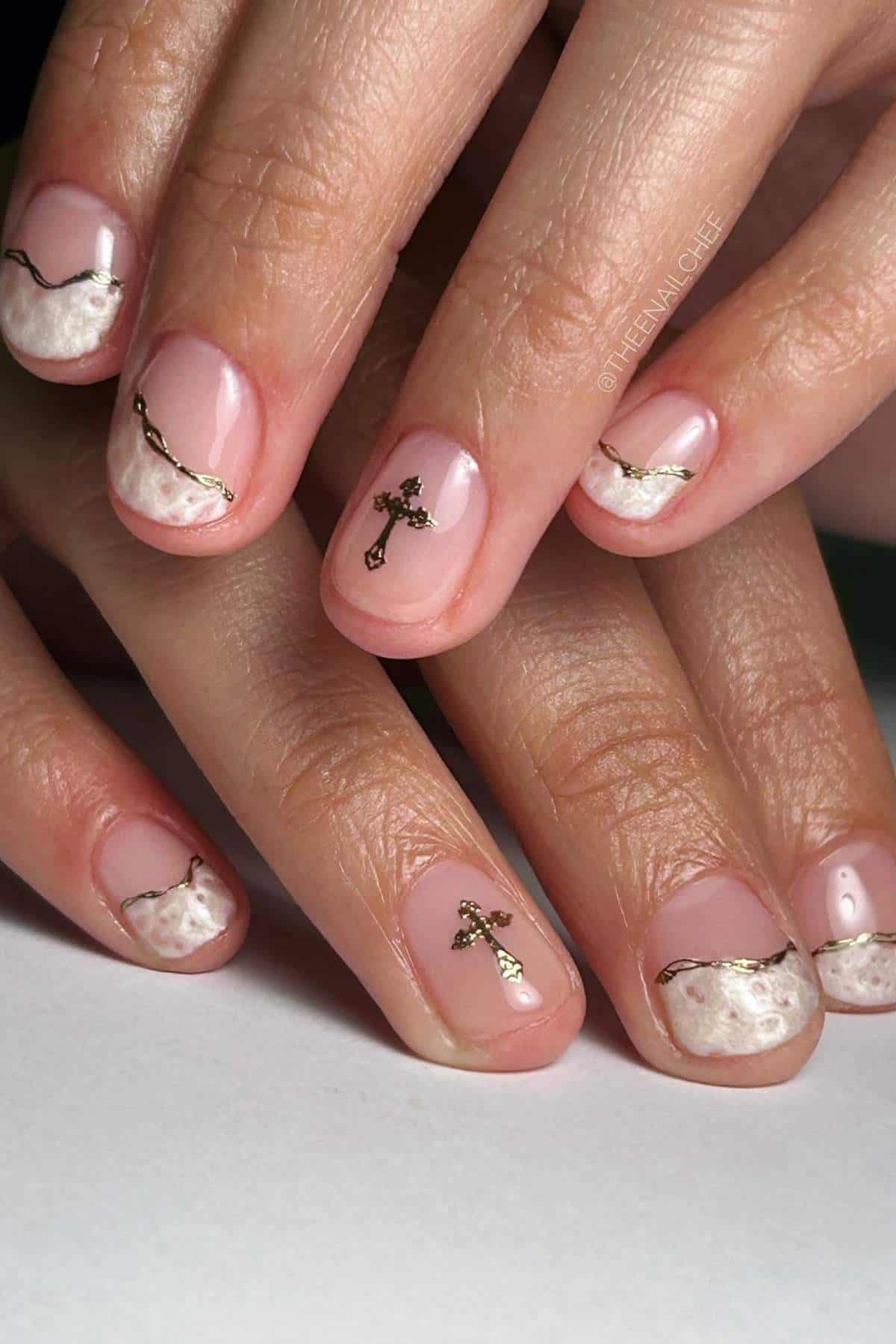 A hand with manicured nails featuring white textured tips, thin gold lines, and small gold cross embellishments on a few nails.