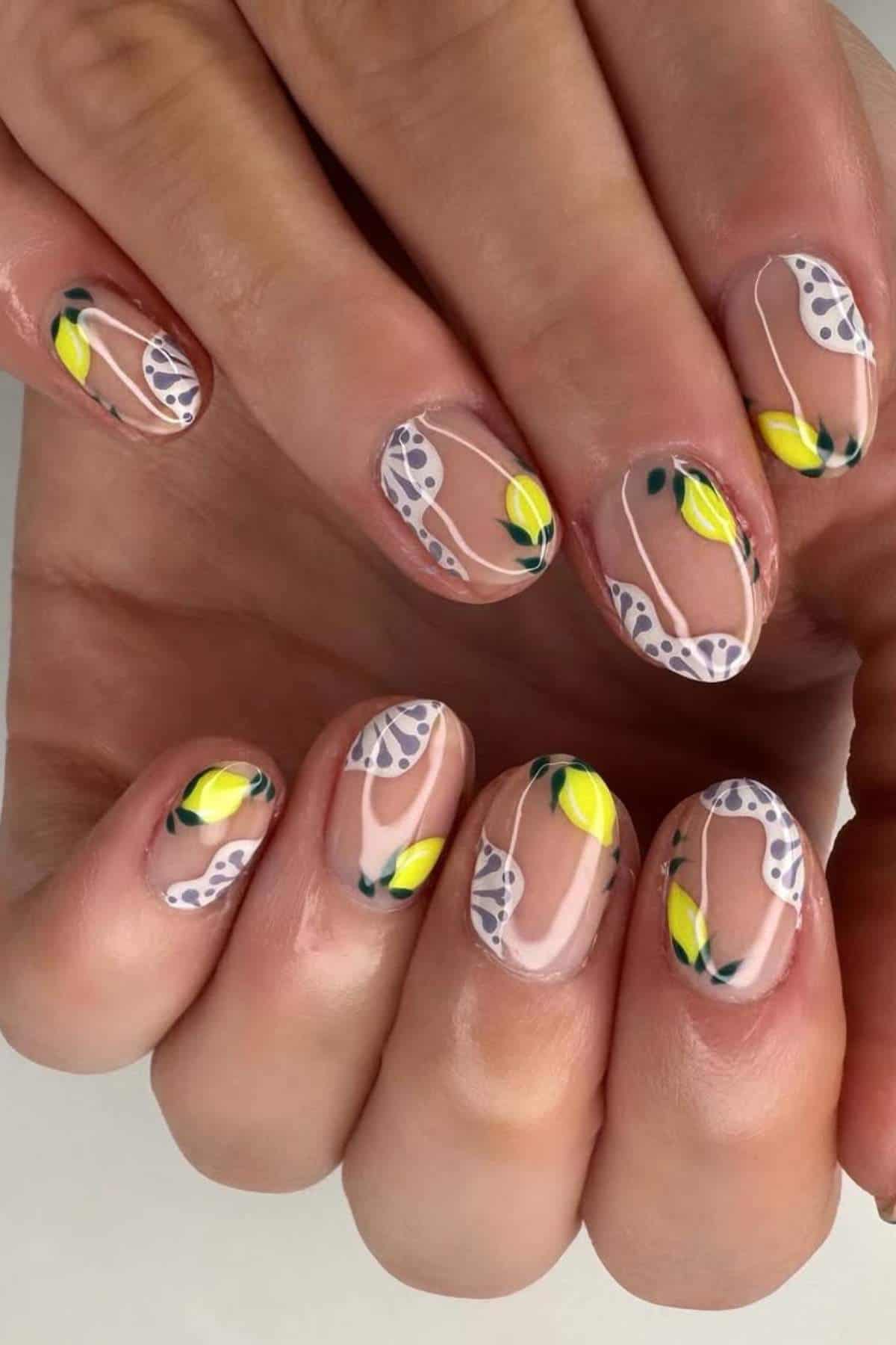 Close-up of hands with short nails featuring clear polish, white abstract shapes, yellow lemon designs, and green leaves as nail art.